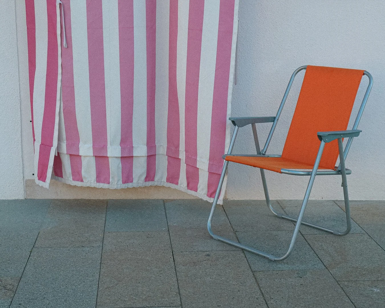 An orange folding chair next to a pink and white striped fabric backdrop on a tiled floor against a white textured wall.