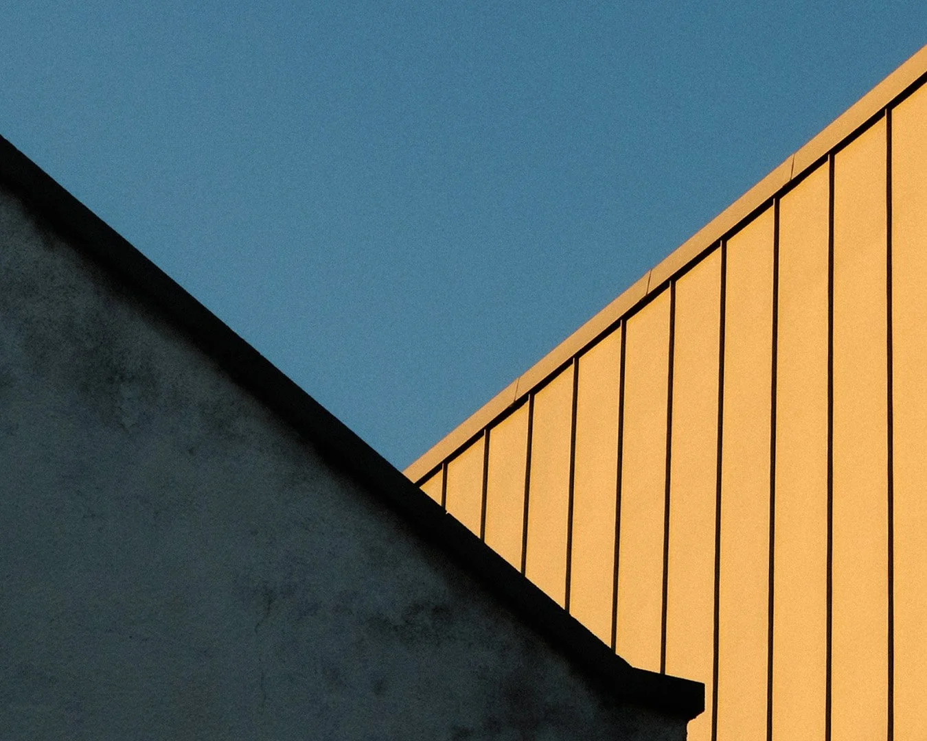 Close-up of building rooftop geometric shapes against a blue sky, featuring a dark textured wall and a beige wall with vertical black lines.