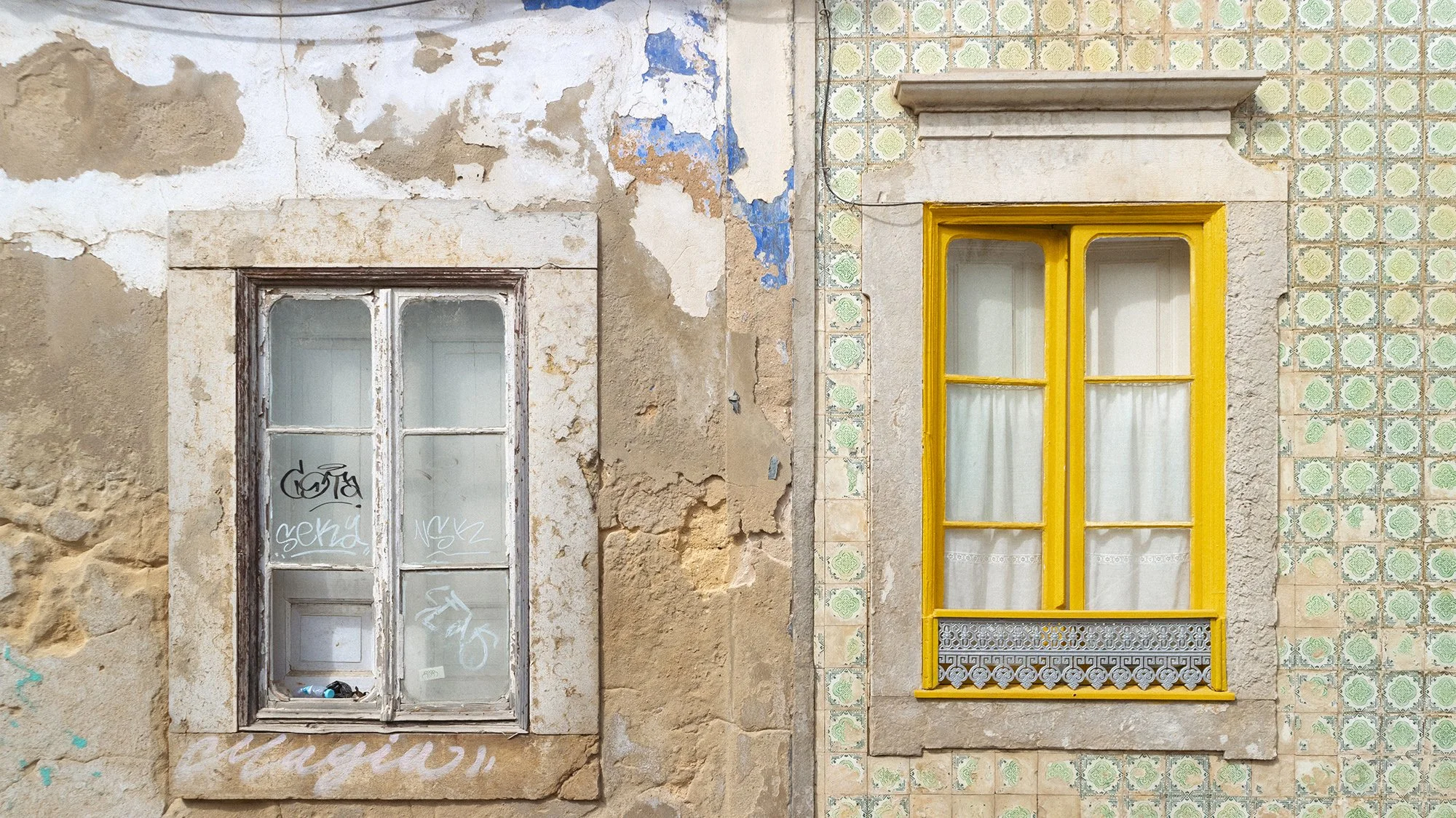 Two windows on a weathered, partially peeling wall with decorative tiles. The window on the left has a white, worn frame with graffiti, while the window on the right has a vibrant yellow frame and a decorative metal grille at the bottom.
