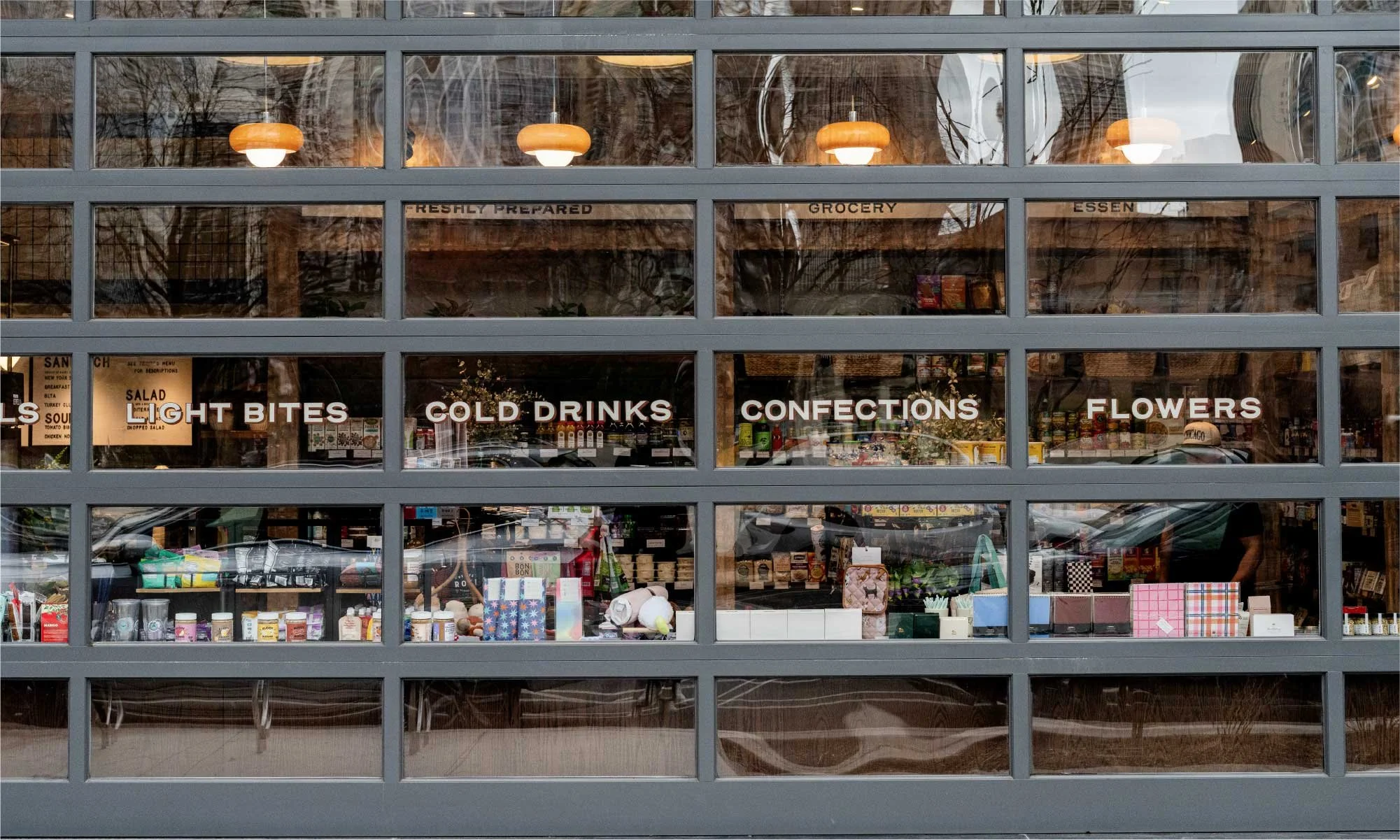 View through a glass window of a store with signs that read "Light Bites," "Cold Drinks," "Confections," and "Flowers." The interior displays various products including packaged goods, flowers, and gifts, with warm lighting and reflections of trees and parked cars outside.