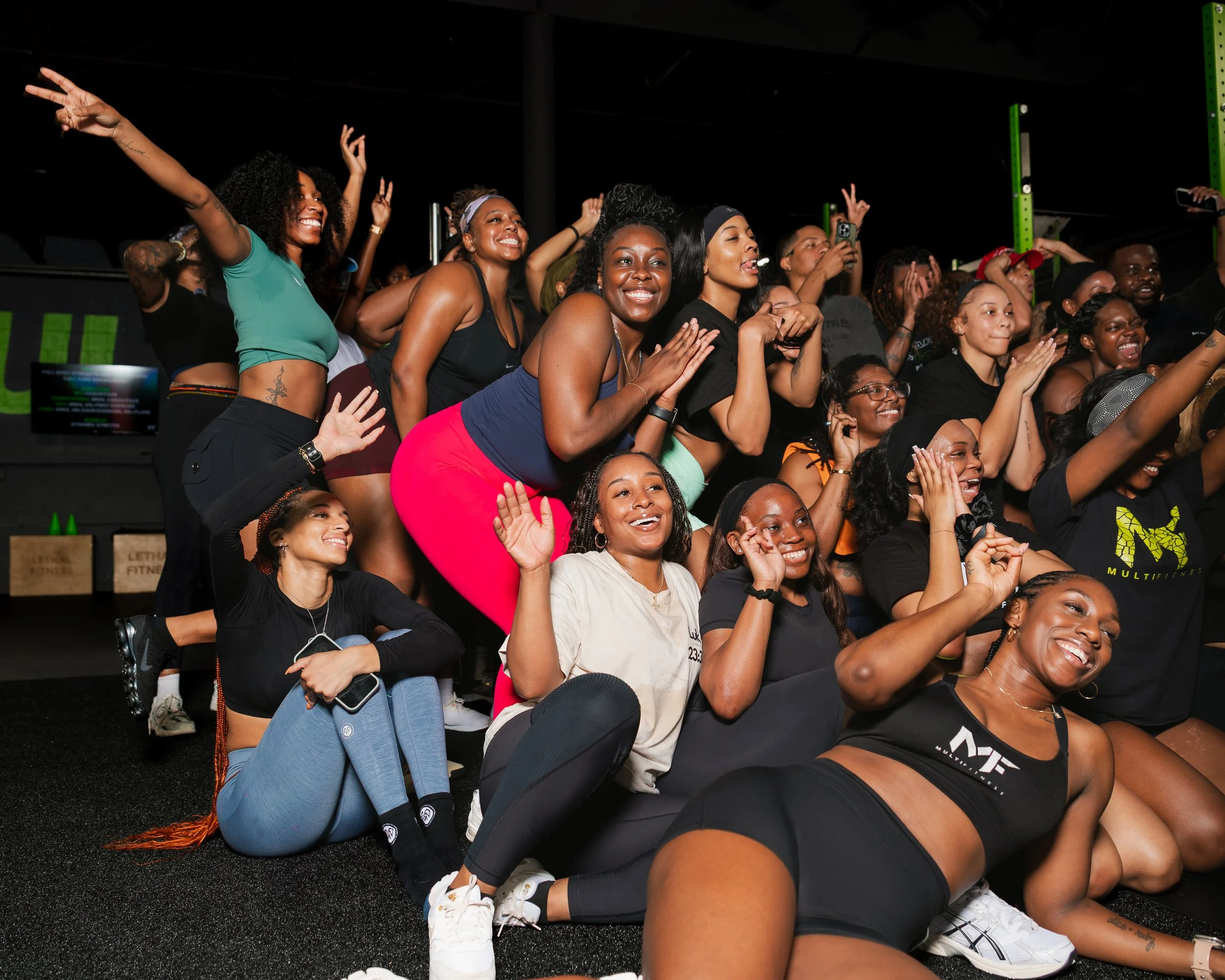 Group of smiling women and girls at a fitness event, some in workout clothes, some taking photos, in a gym or fitness center.