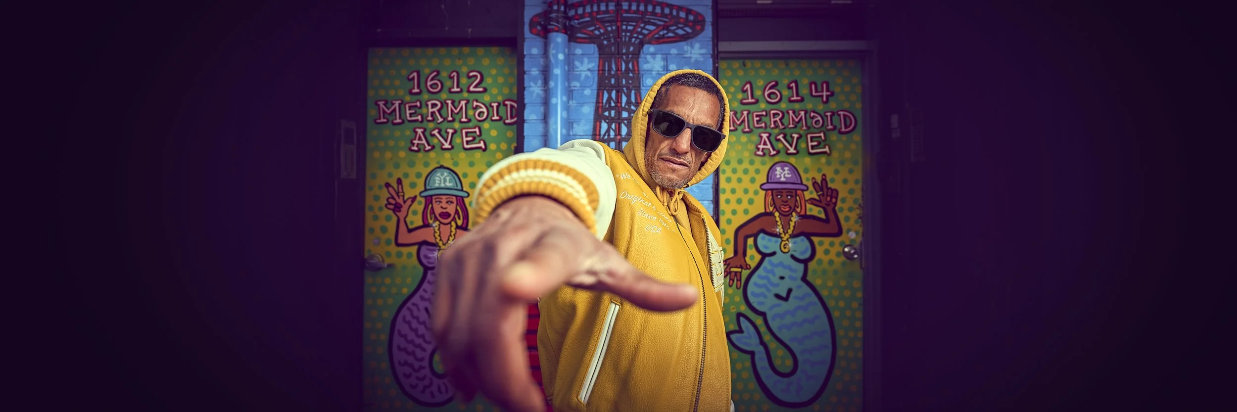 A man wearing sunglasses, a yellow hoodie, and a yellow jacket pointing towards the camera with a serious expression. Behind him are colorful murals of mermaids and signs reading '1612 Mermaid Ave' and '1614 Mermaid Ave'.