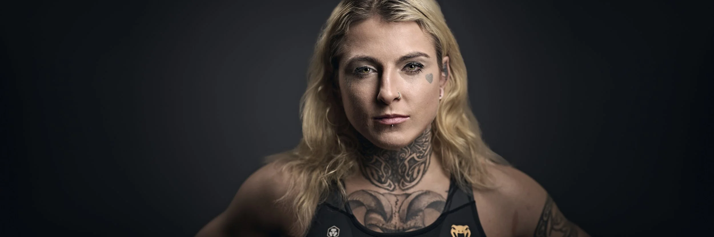 Close-up of a woman with blonde wavy hair and tattoos, wearing a black sports top, against a dark background.