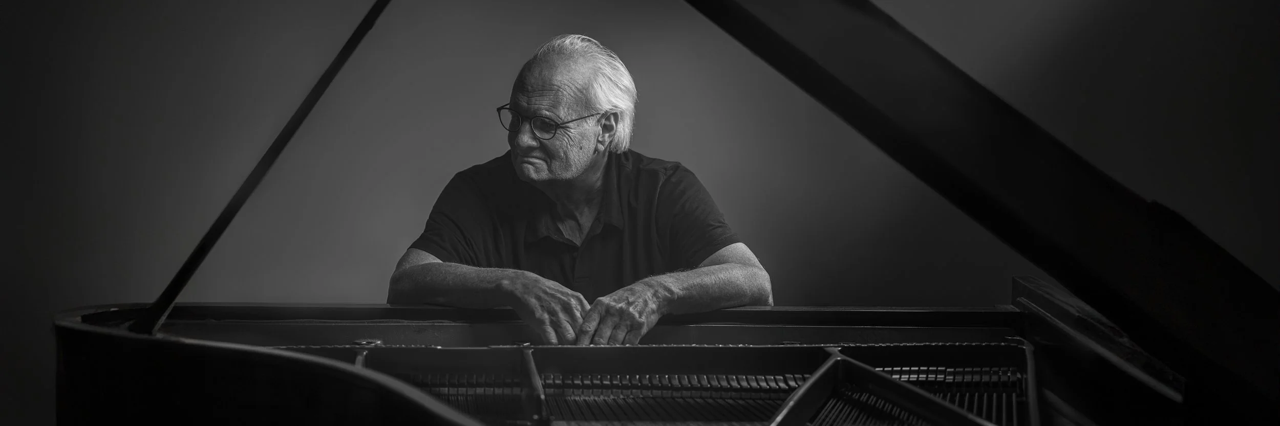 Black and white photo of an elderly man with glasses and white hair, sitting at a grand piano with his arms resting on the piano's open lid, looking contemplative.