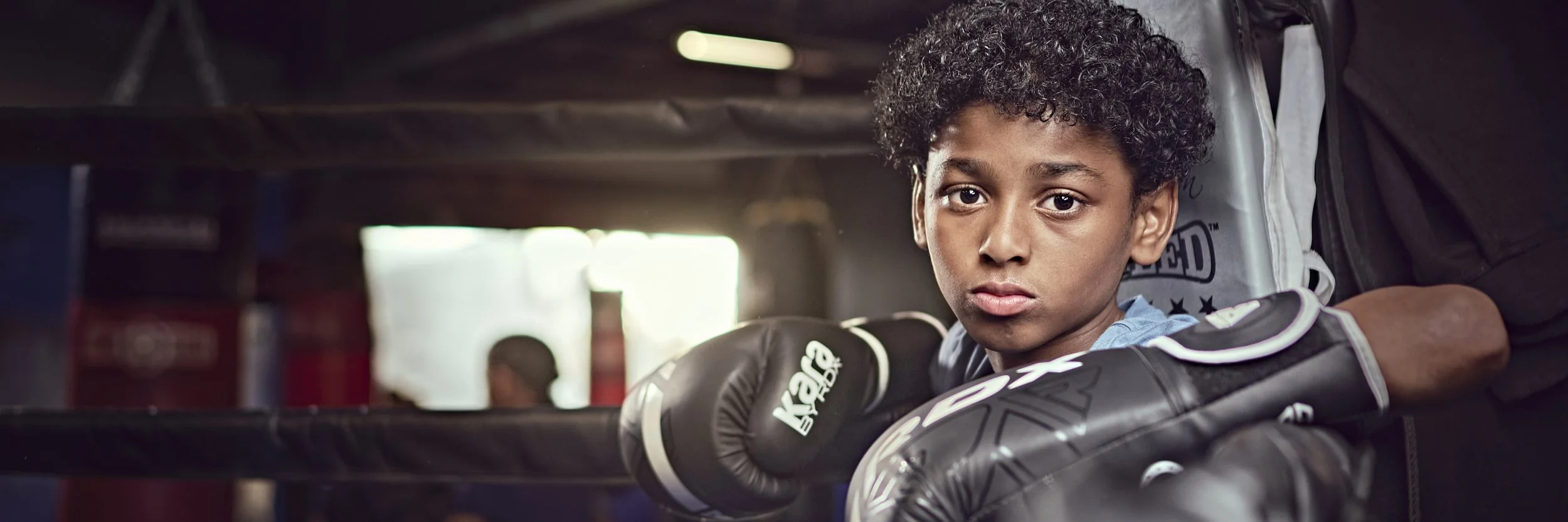 Young boy with curly hair wearing boxing gloves resting his arms on a boxing ring's corner pad in a gym.