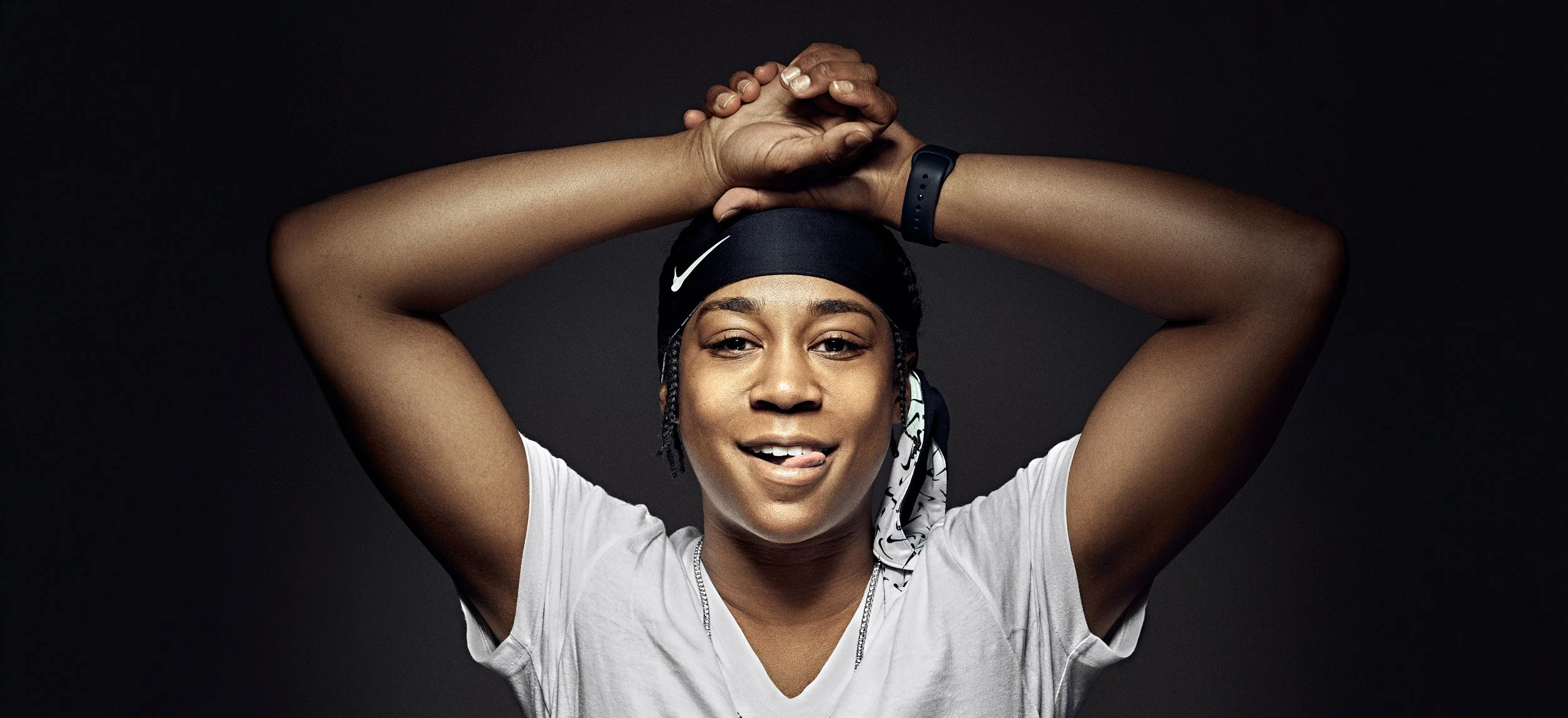 A woman with braided hair, wearing a black headband, white T-shirt, and a black watch, smiling with tongue slightly out, against a dark background.