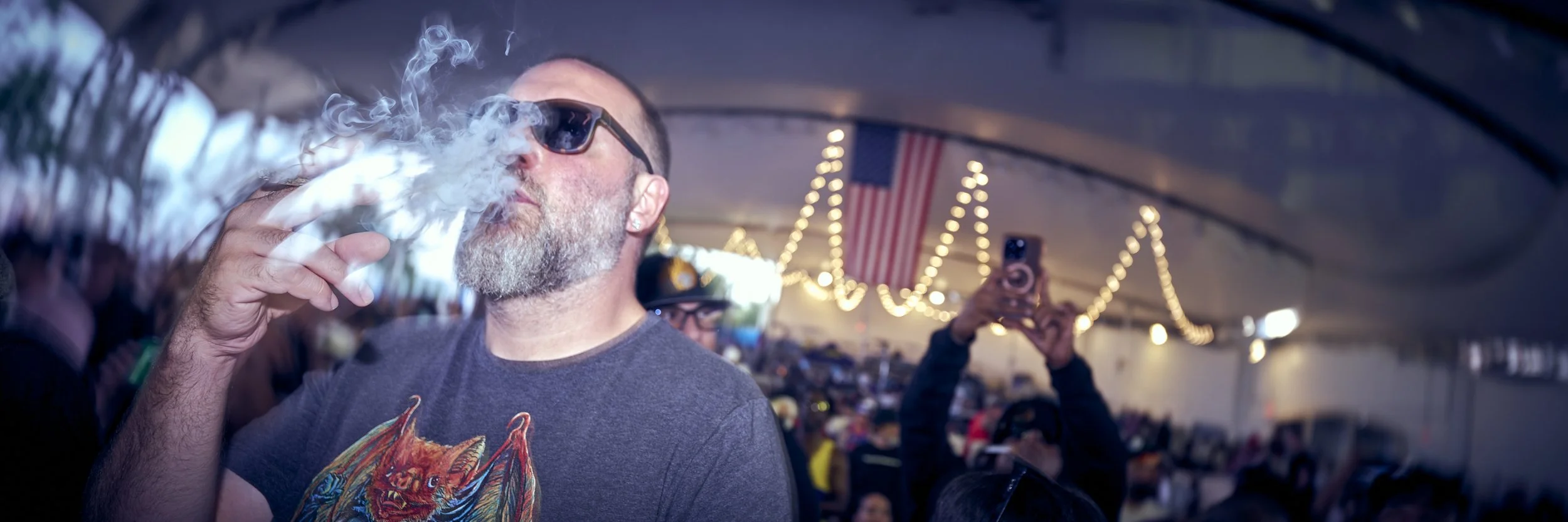 A man with a beard and sunglasses exhaling smoke in a crowded event with string lights and American flags in the background.