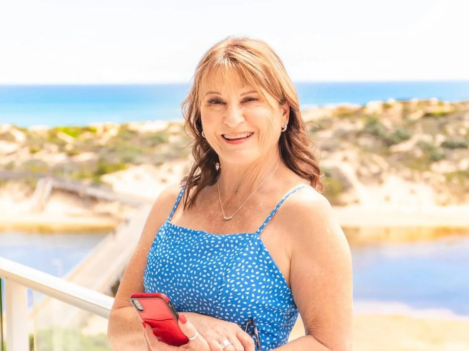 A smiling woman with shoulder-length red hair wearing a blue sleeveless dress with white polka dots, holding a red smartphone, with a backdrop of a beach and ocean.