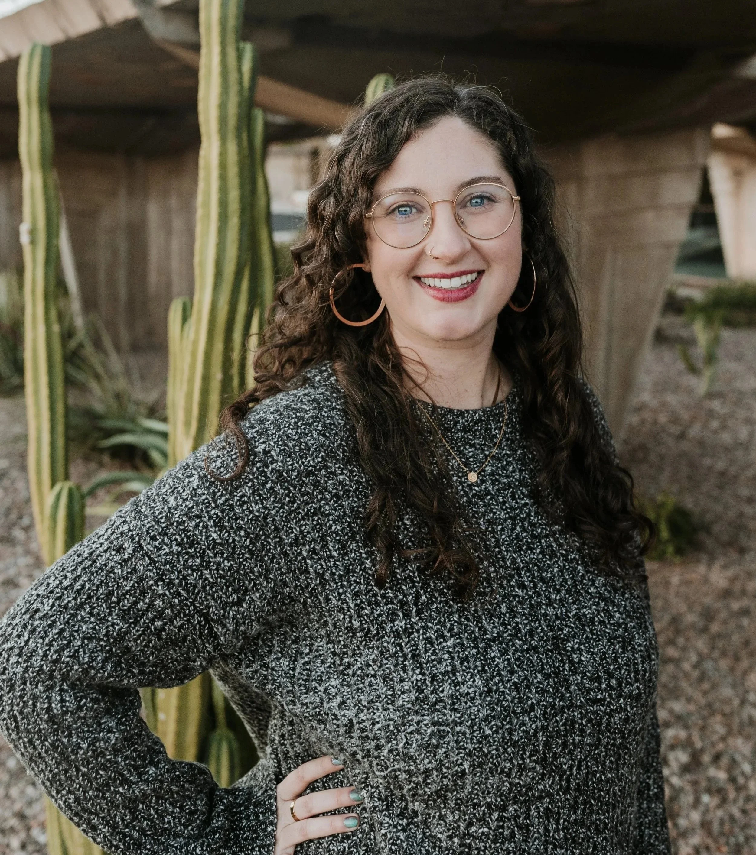 A woman with curly brown hair and blue eyes wearing glasses, earrings, and a patterned sweater, standing outdoors near a cactus plant and a wooden structure.
