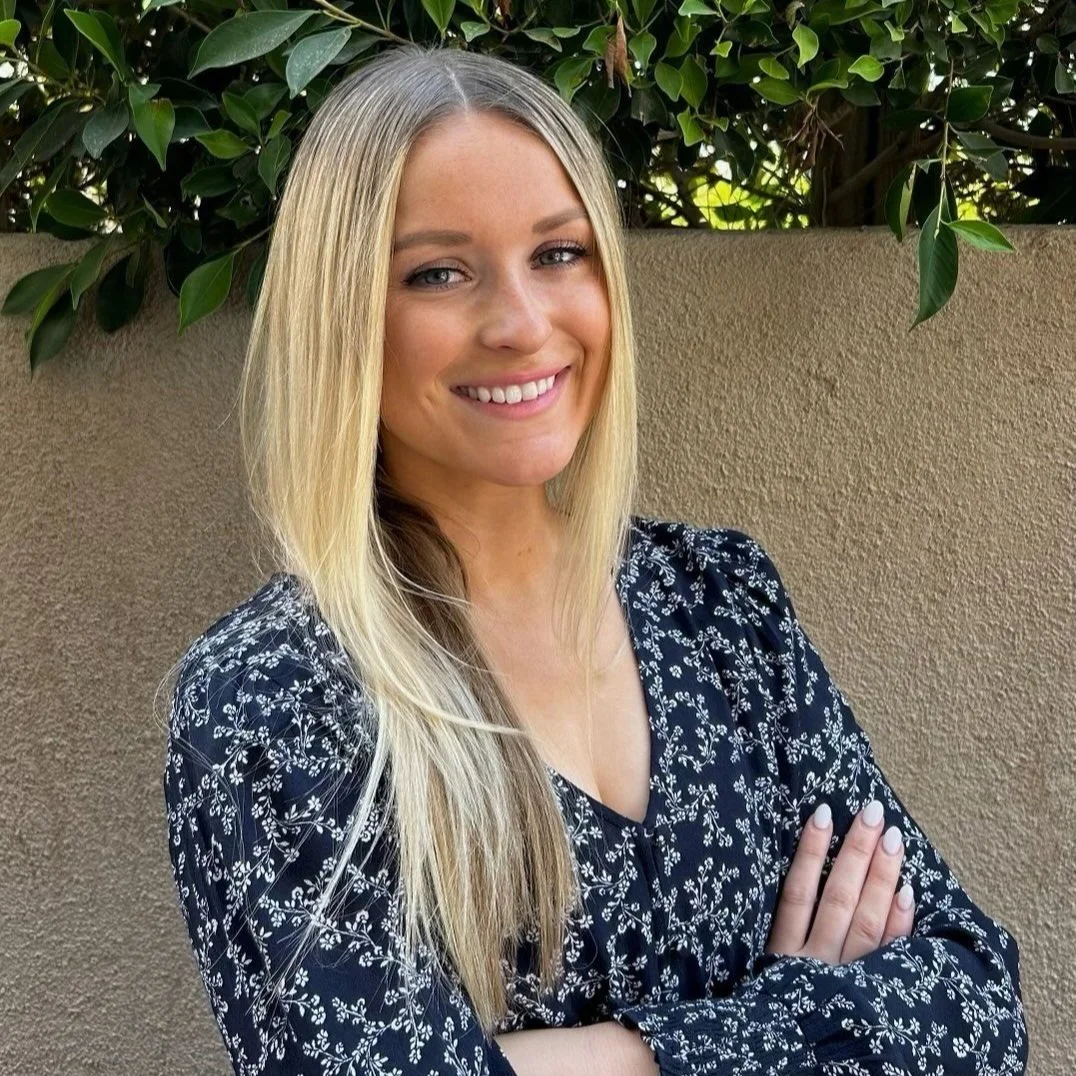 A young woman with long blonde hair and blue eyes smiling, standing with arms crossed in front of a beige wall with green foliage overhead.