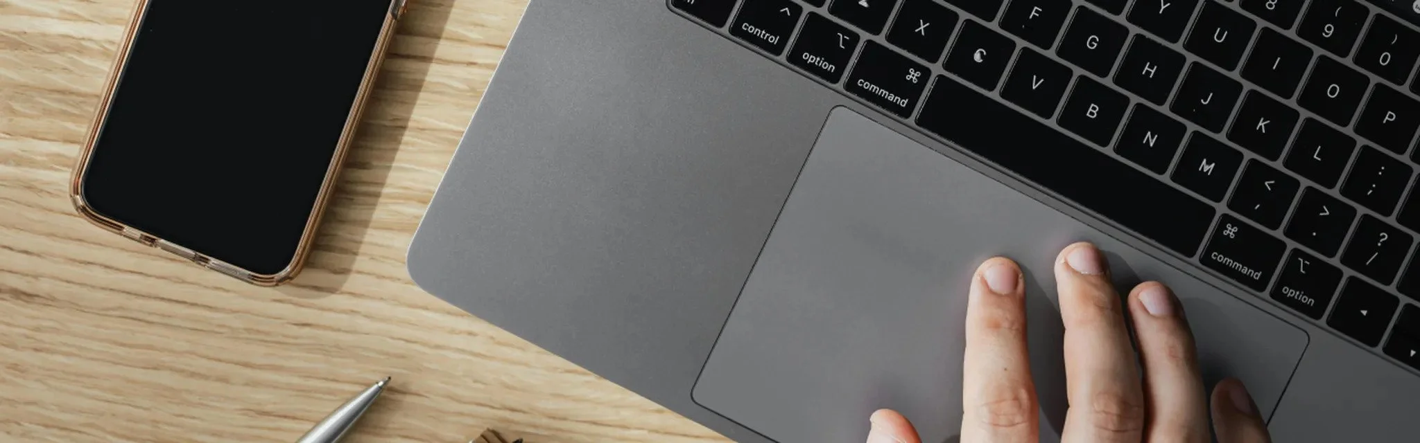 A hand using a gray laptop touchpad with a smartphone placed nearby on a light wooden desk surface. A pen is also visible in the workspace.