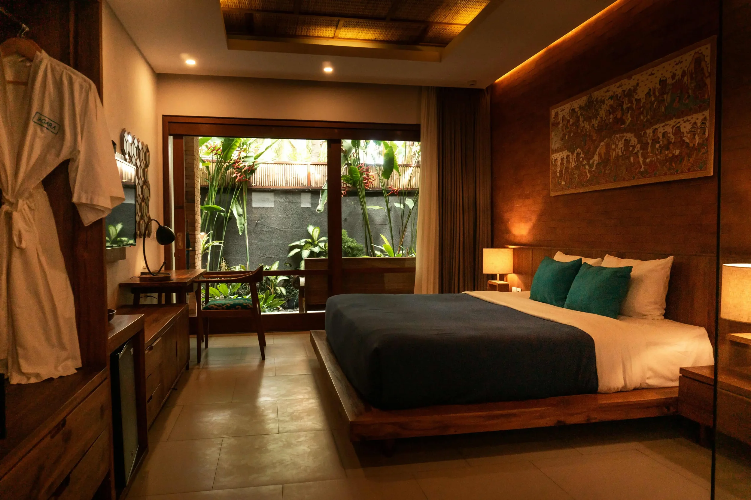 A modern hotel room with warm wood furnishings and large windows overlooking a lush tropical courtyard.