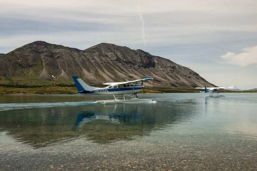 Two seaplanes landing on a clear lake with mountains in the background under a partly cloudy sky.