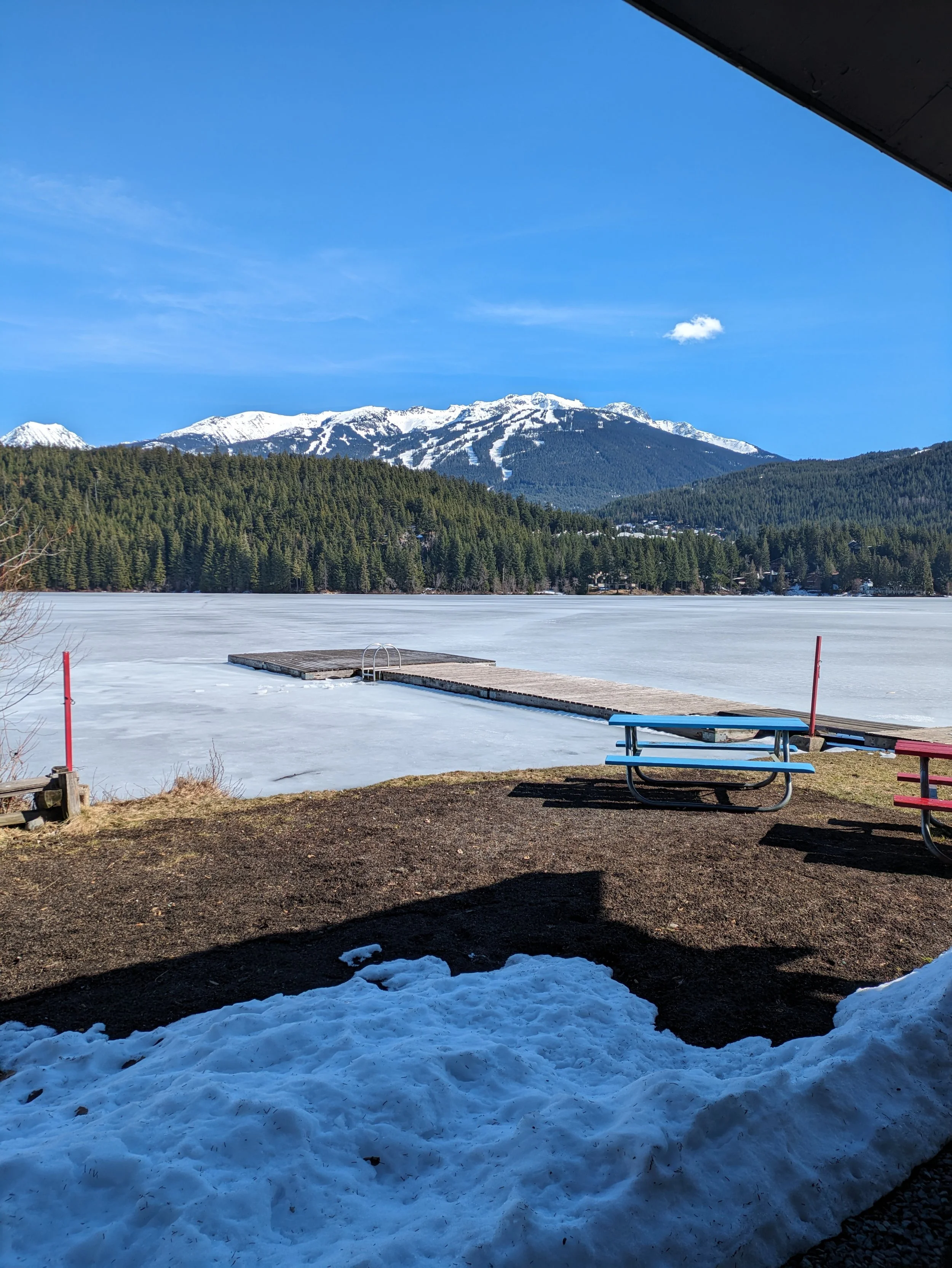 Photo of the lake, frozen, with the dock in the foreground and mountains in the back.