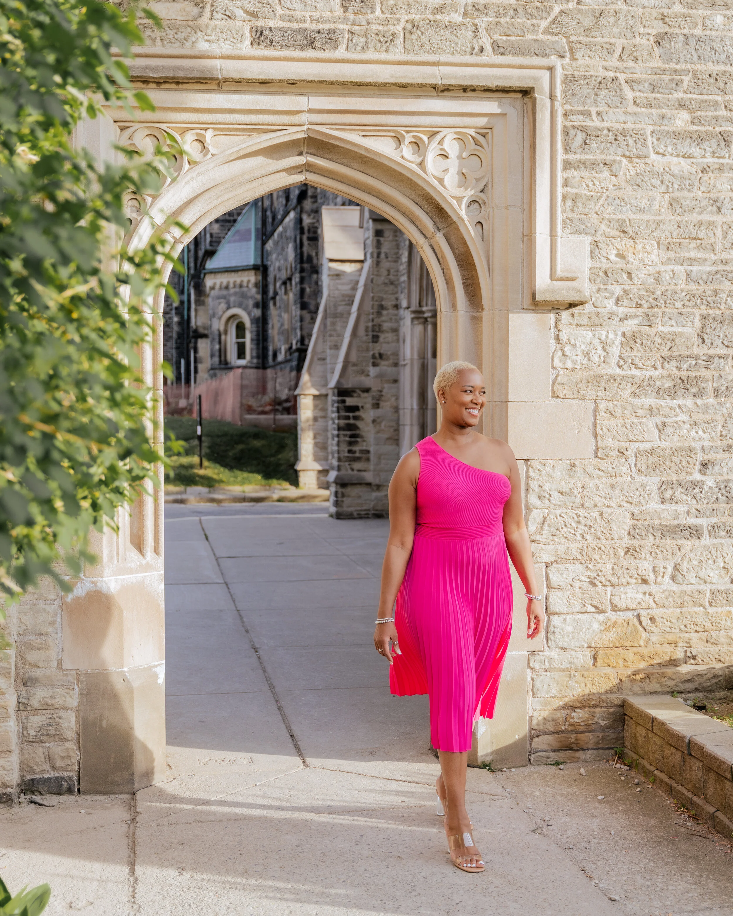 A woman wearing a bright pink, asymmetrical dress with pleats, walking through an arched stone gateway of a historic building, smiling and looking to the right.
