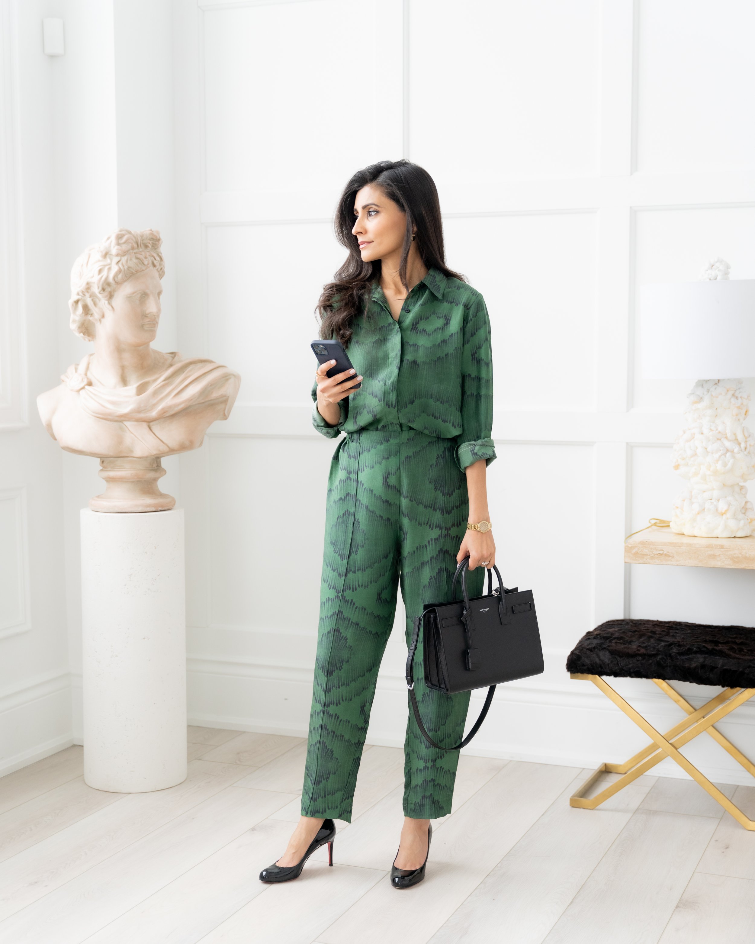 A woman in a green patterned suit stands indoors with a bust sculpture to her left, holding a smartphone in one hand and a black handbag in the other, wearing black high heels.