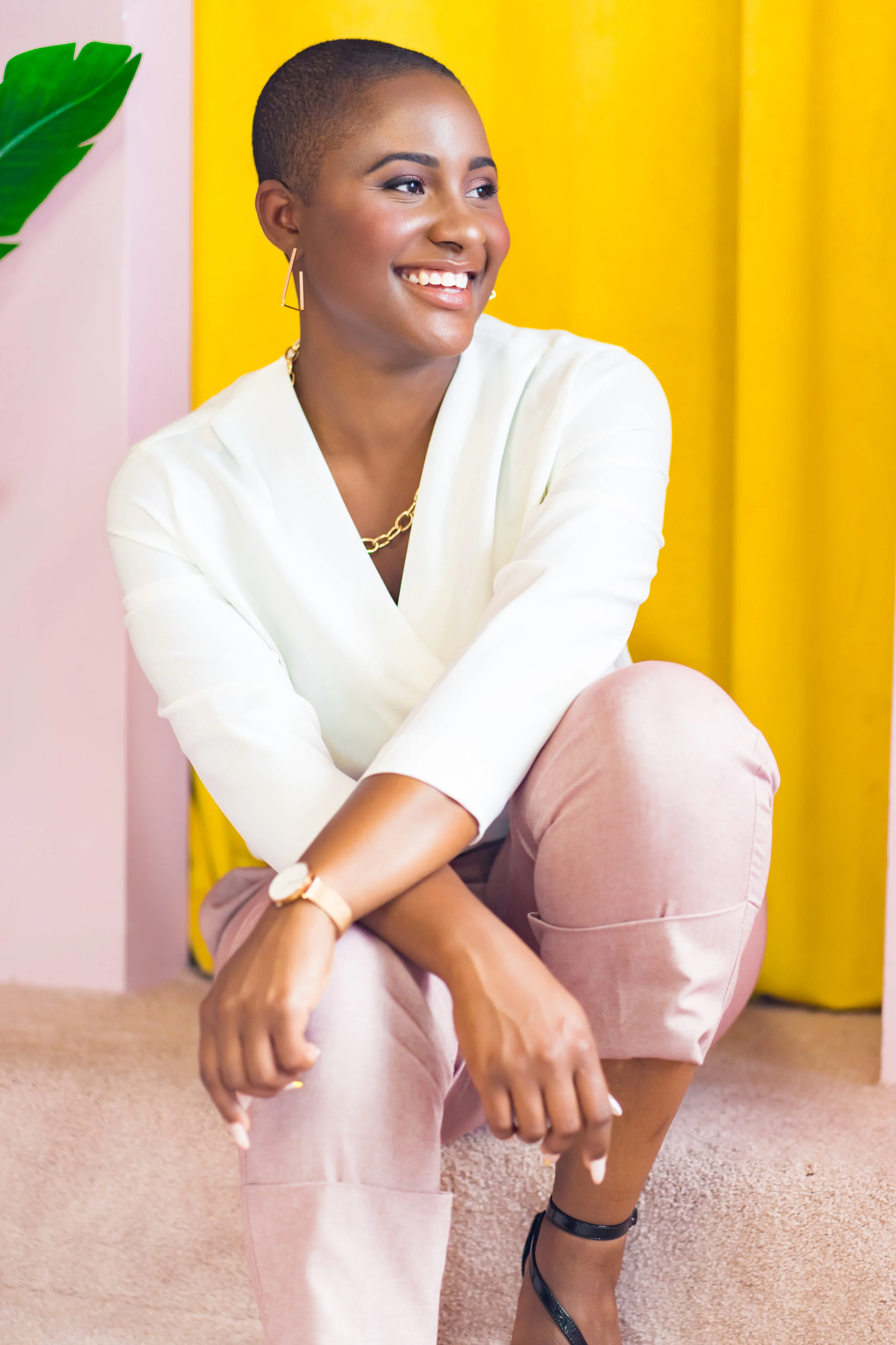 A smiling woman with short hair, wearing a white blouse, pink pants, and black heels, sitting on the floor against a colorful background of pink, yellow, and green.