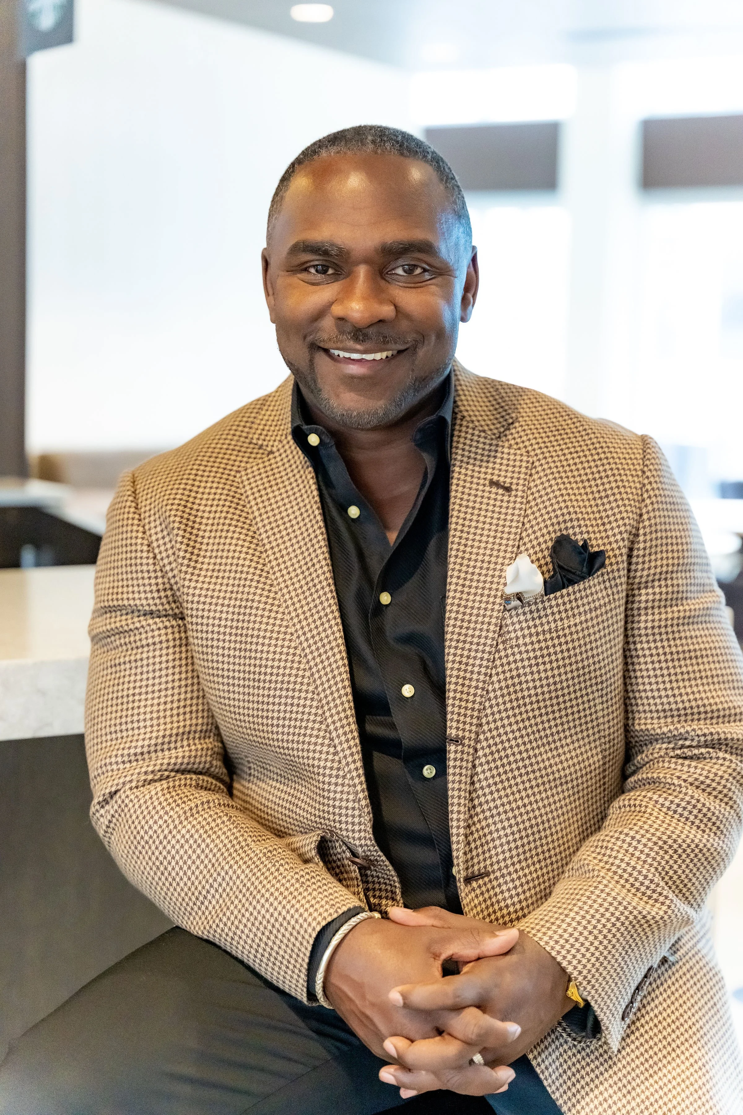 A smiling man wearing a beige houndstooth blazer, black shirt, and pocket square, sitting in a modern indoor setting.