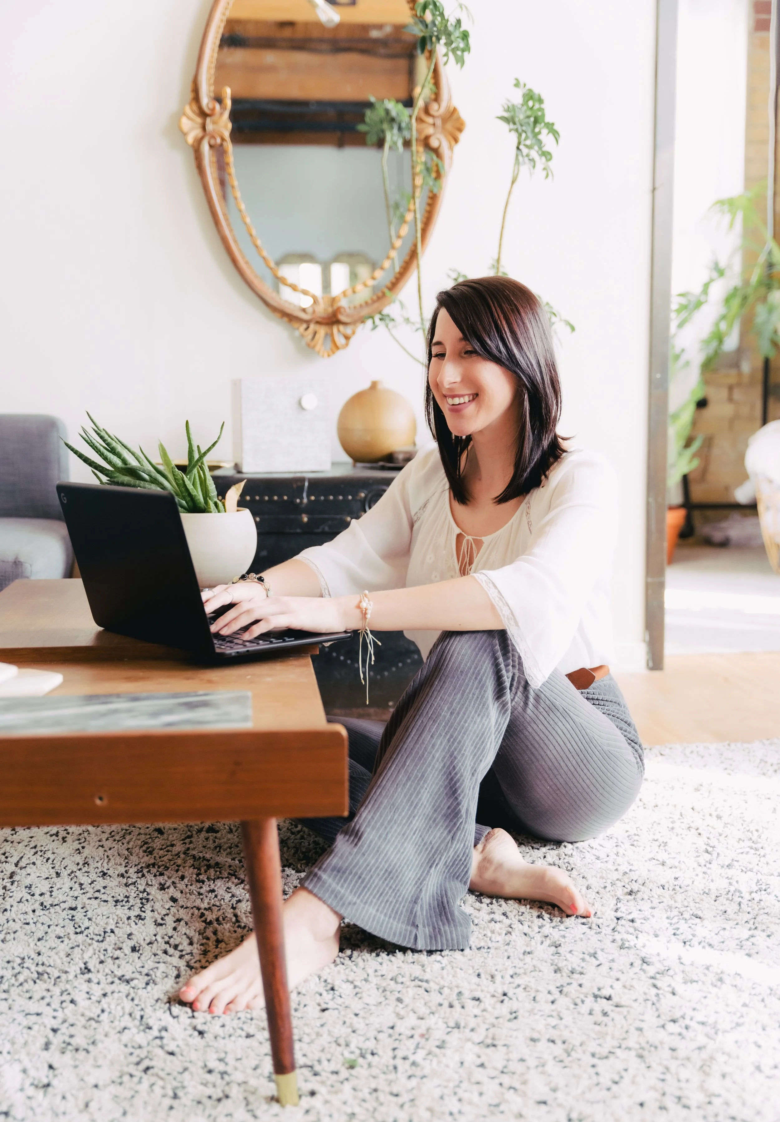 A woman with dark hair sitting on a cozy rug next to a wooden coffee table, working on a laptop, smiling, in a brightly lit living room with plants, a mirror, and decorative items.