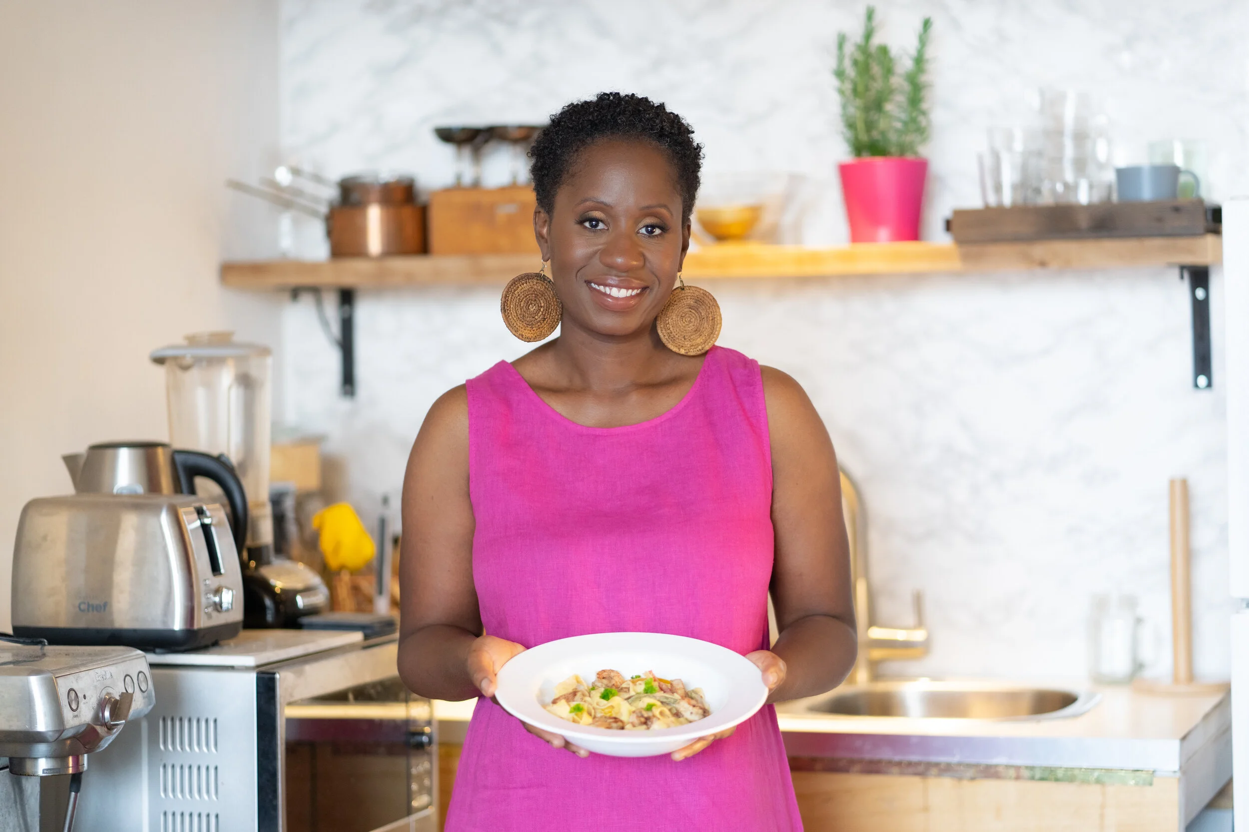 A woman in a pink sleeveless dress holding a bowl of pasta in a kitchen with a brick wall background.
