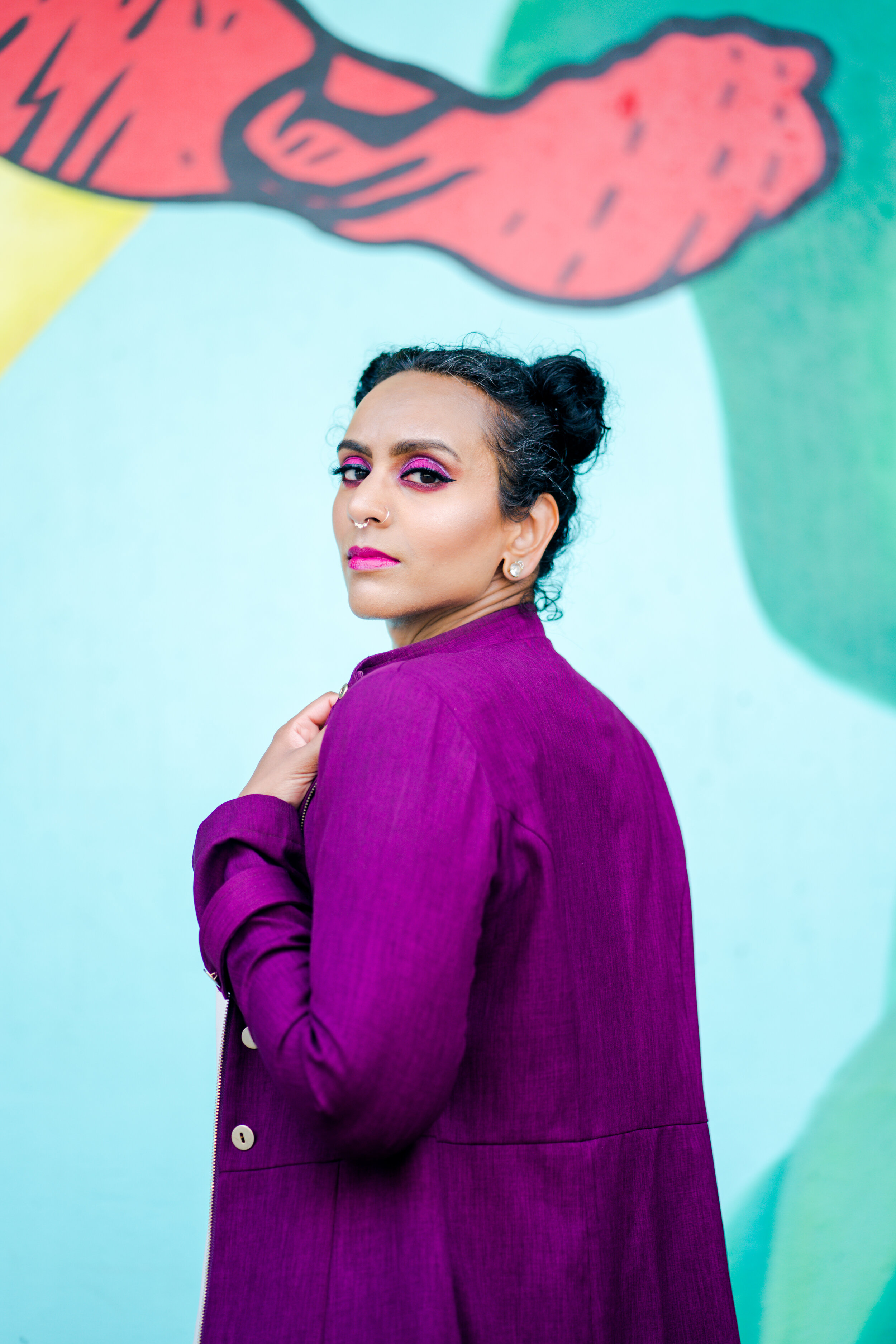 Woman with styled hair and bright makeup wearing a purple jacket, standing in front of a colorful mural on the wall.