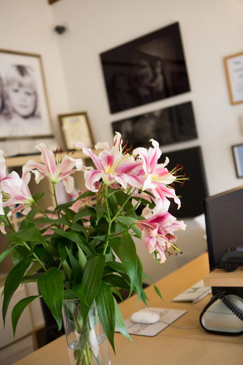 Pink and white lilies in a glass vase on a desk in an office. In the background, there are framed pictures and awards on the wall, and a computer monitor and phone on the desk.