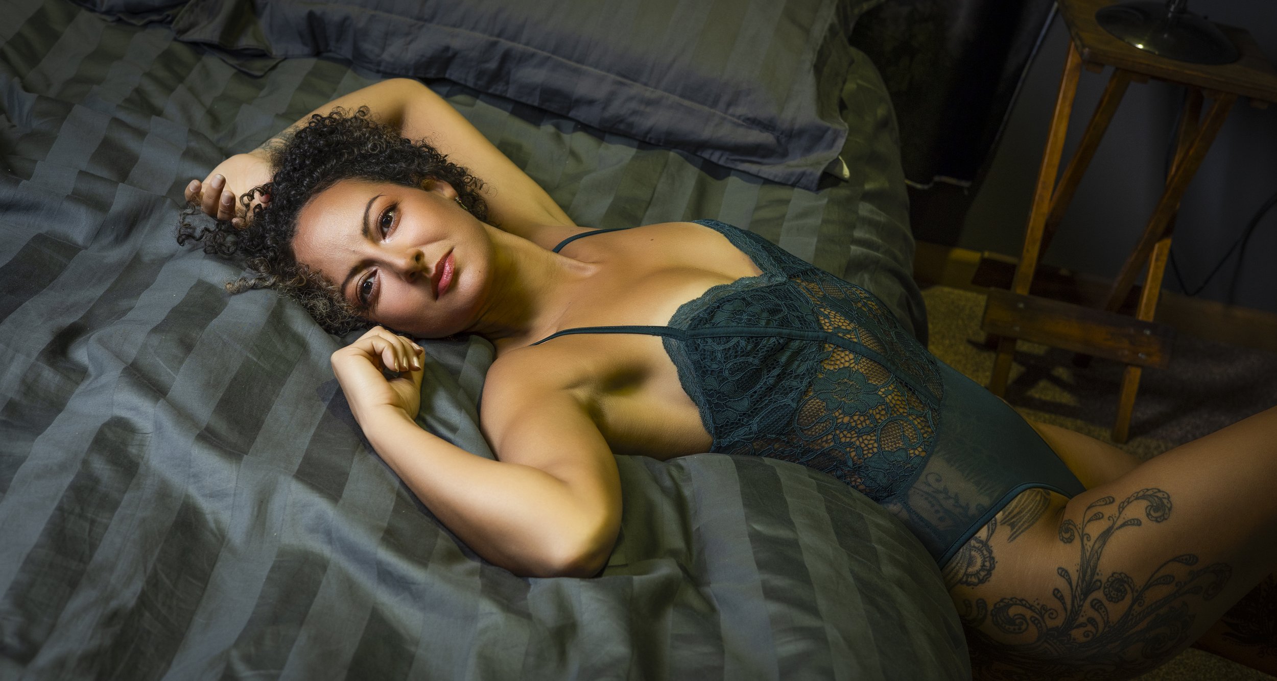 A woman with curly hair lying on a bed, wearing a dark green lace lingerie, looking at the camera, with tattoos on her right arm and thigh.