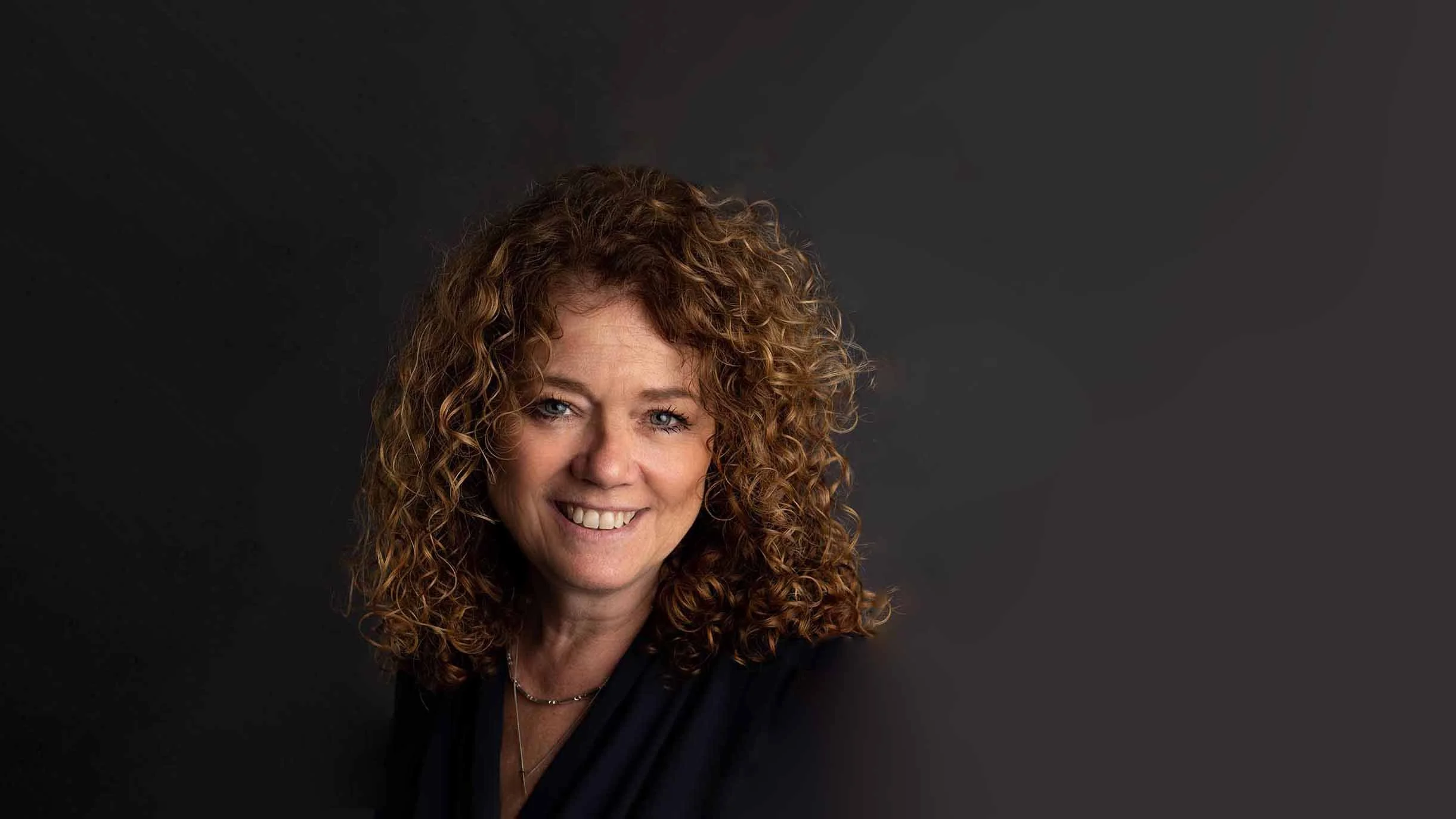 A woman with curly red hair smiling against a dark background.