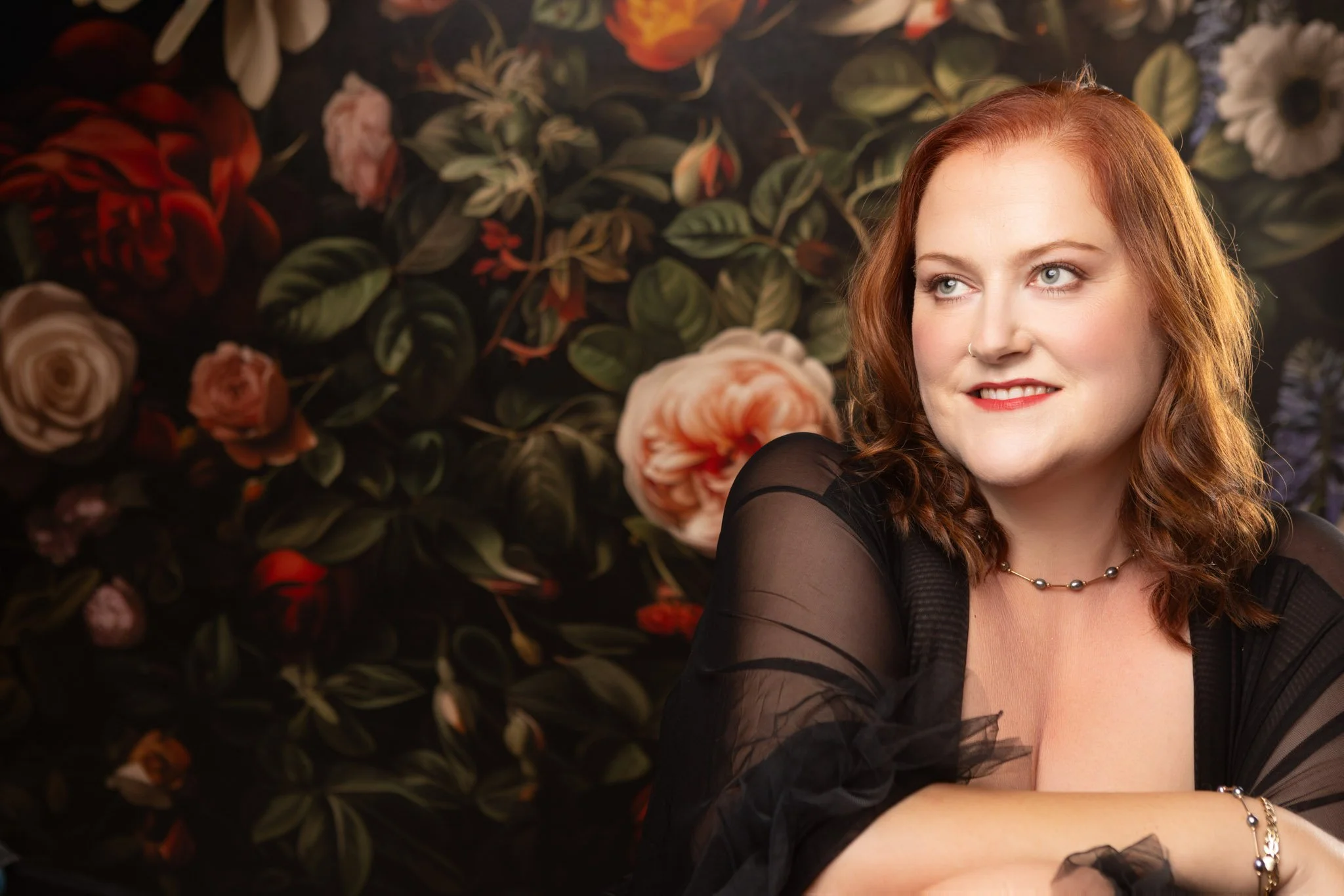 A woman with red hair and light-colored eyes posing in front of a dark floral background, wearing an off-shoulder black top and jewelry.