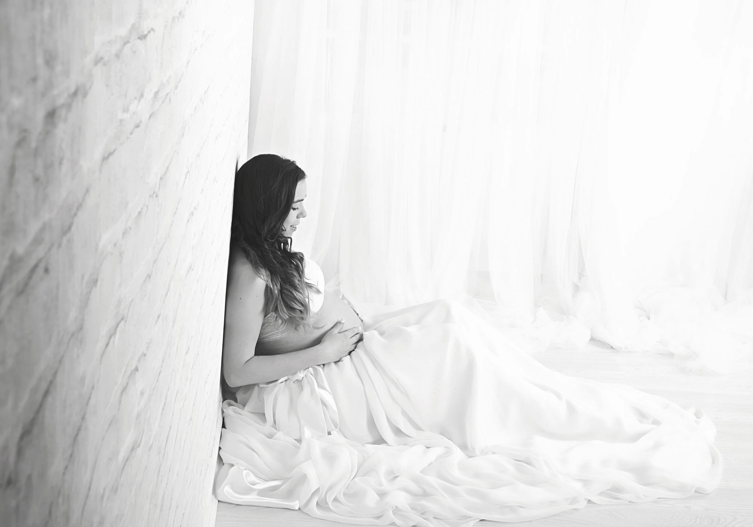A pregnant woman sitting on the floor, leaning against a wall, wearing a flowing white gown, with her eyes closed and hands resting on her belly, in a bright, softly lit room with sheer curtains.