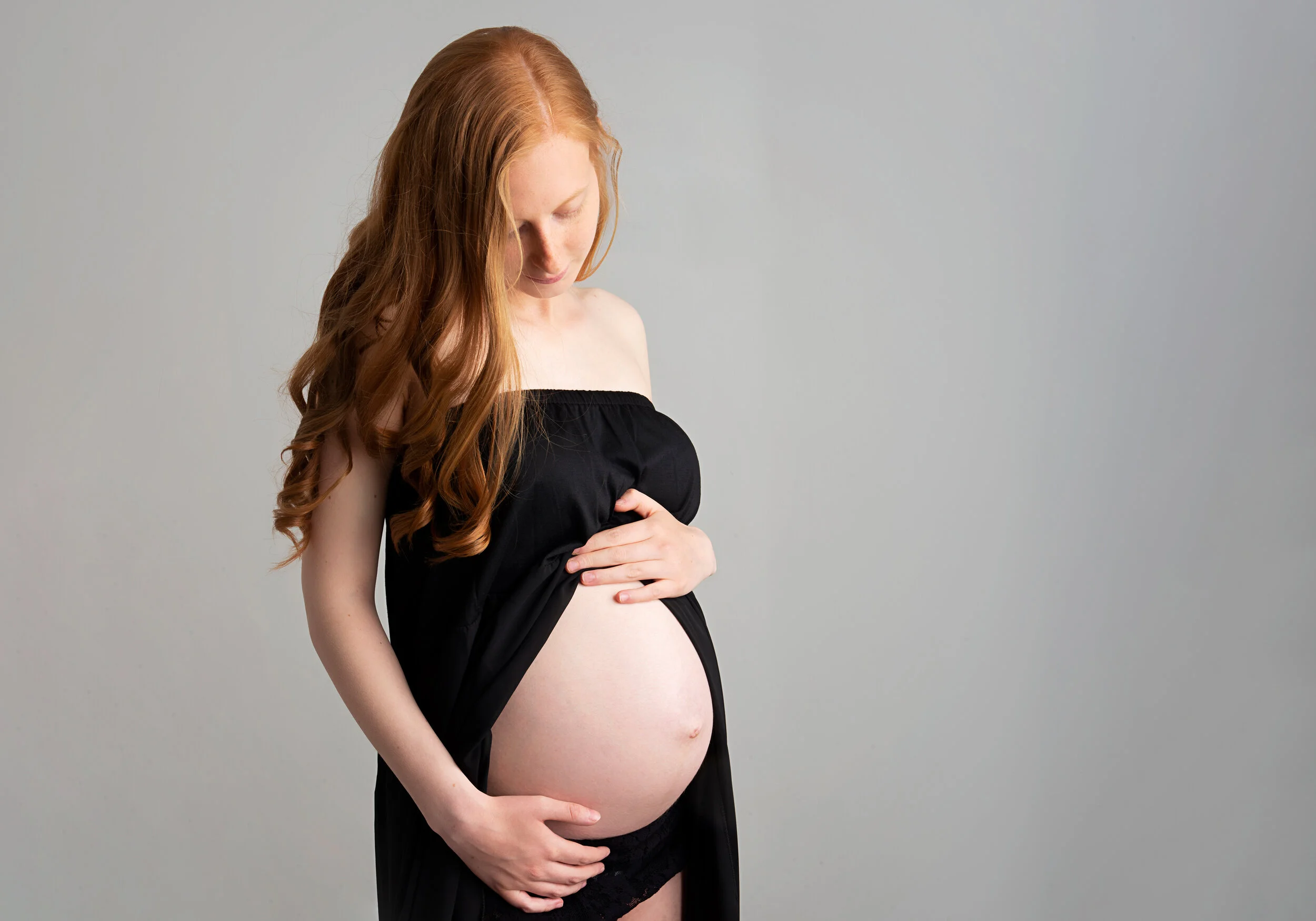 A pregnant woman with long red hair, wearing a black strapless top and black lace underwear, gently holding her baby bump while looking down at it against a plain gray background.