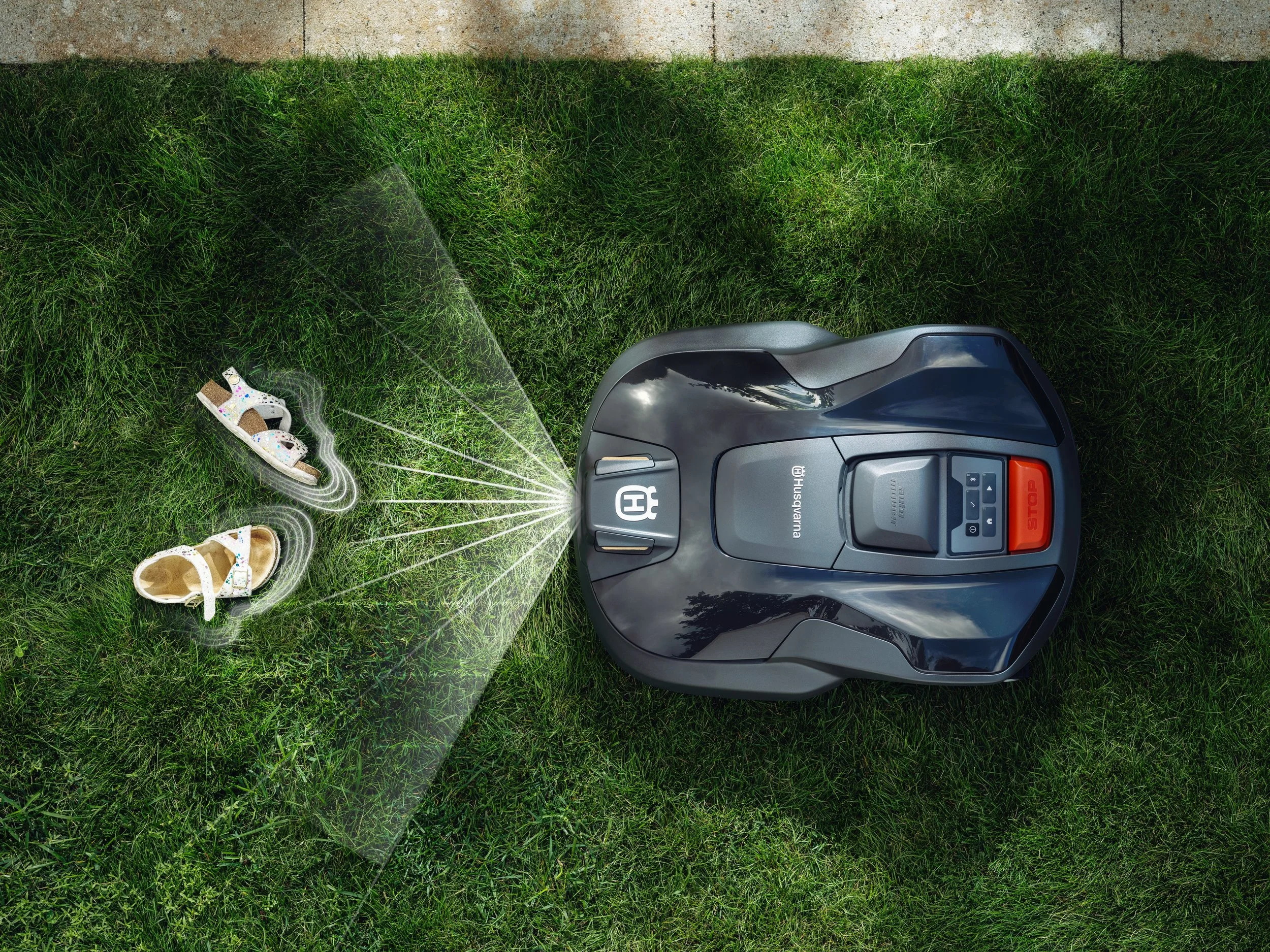 A robotic lawn mower with a black and gray body mowing the grass, with two pairs of children's shoes nearby on the grass.