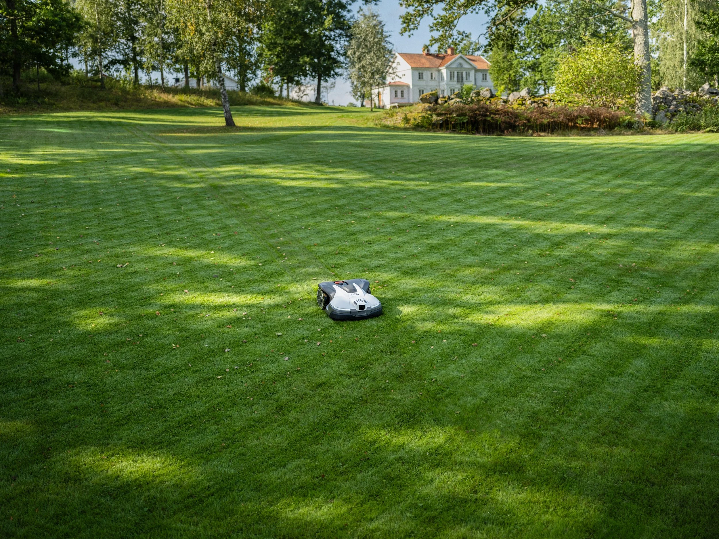 A robotic lawn mower on a well-manicured green lawn with trees and a large house in the background.