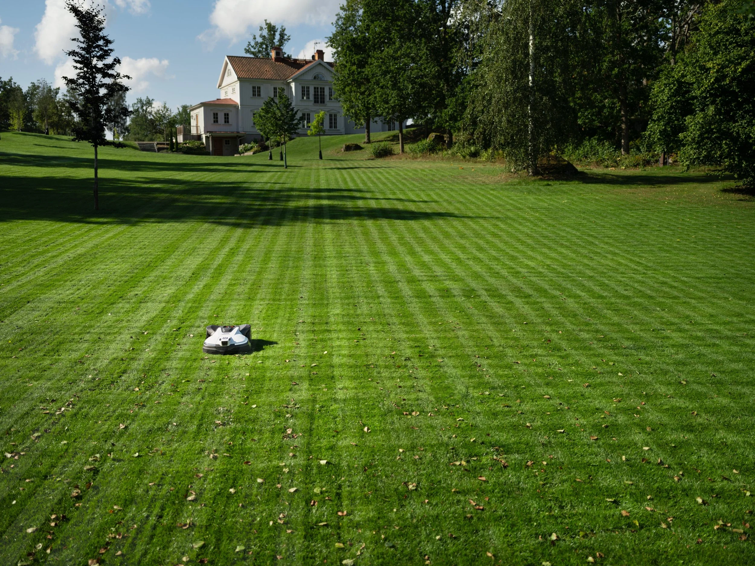 A robotic lawn mower operating on a large, well-maintained lawn with striped grass, trees, and a white house in the background.