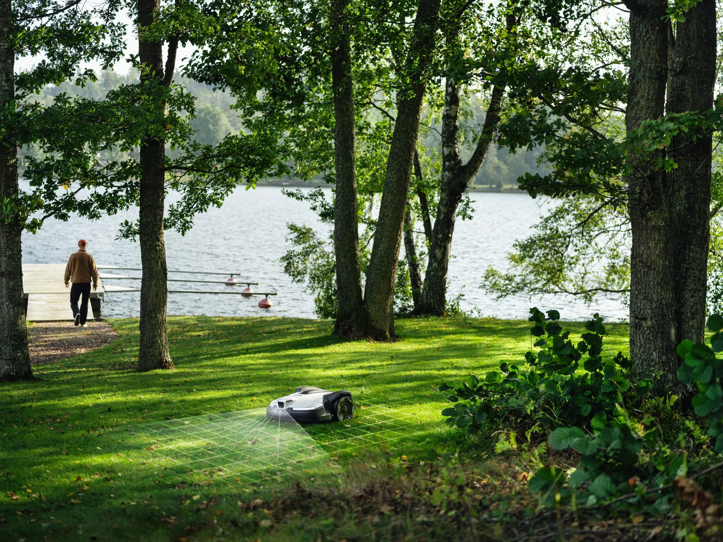A robotic lawn mower working on a grassy area near a lake, with a person walking on a dock in the background surrounded by trees.