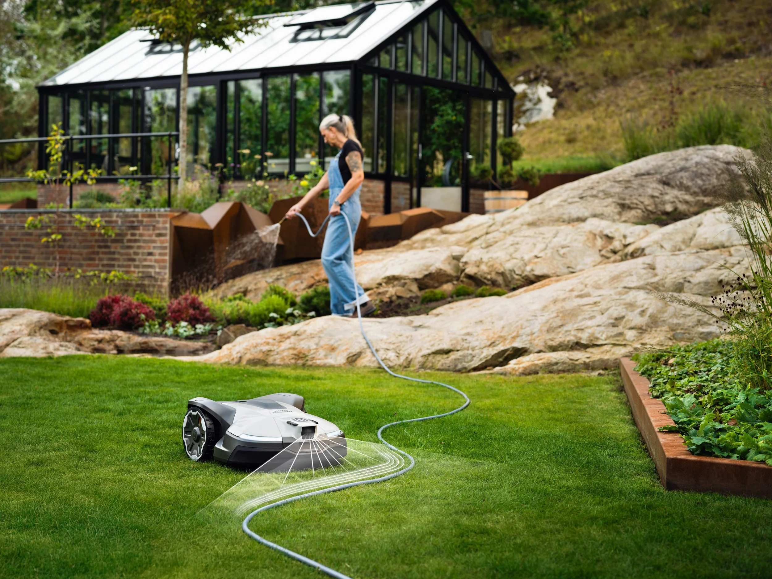 A woman with gray hair, tattoos, wearing a black shirt and blue overalls, is watering plants in a garden using a garden hose connected to a robotic lawn mower on the grass.