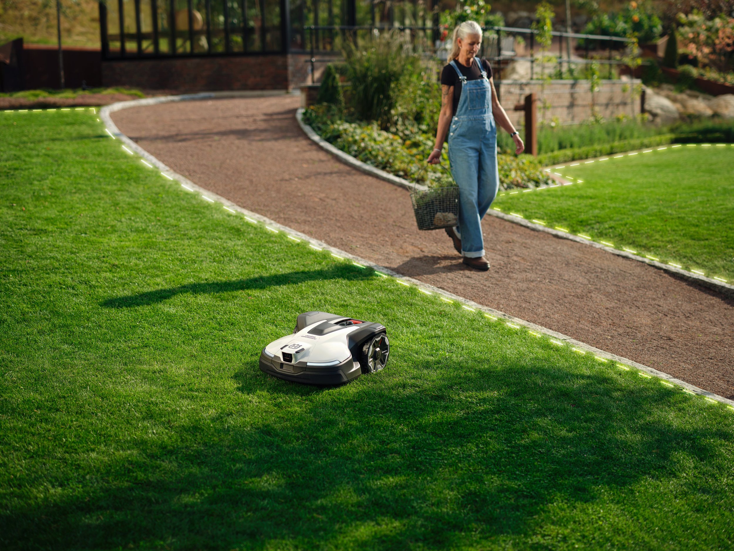 A woman walking on a garden path in a yard with a robotic lawn mower cutting the grass nearby.