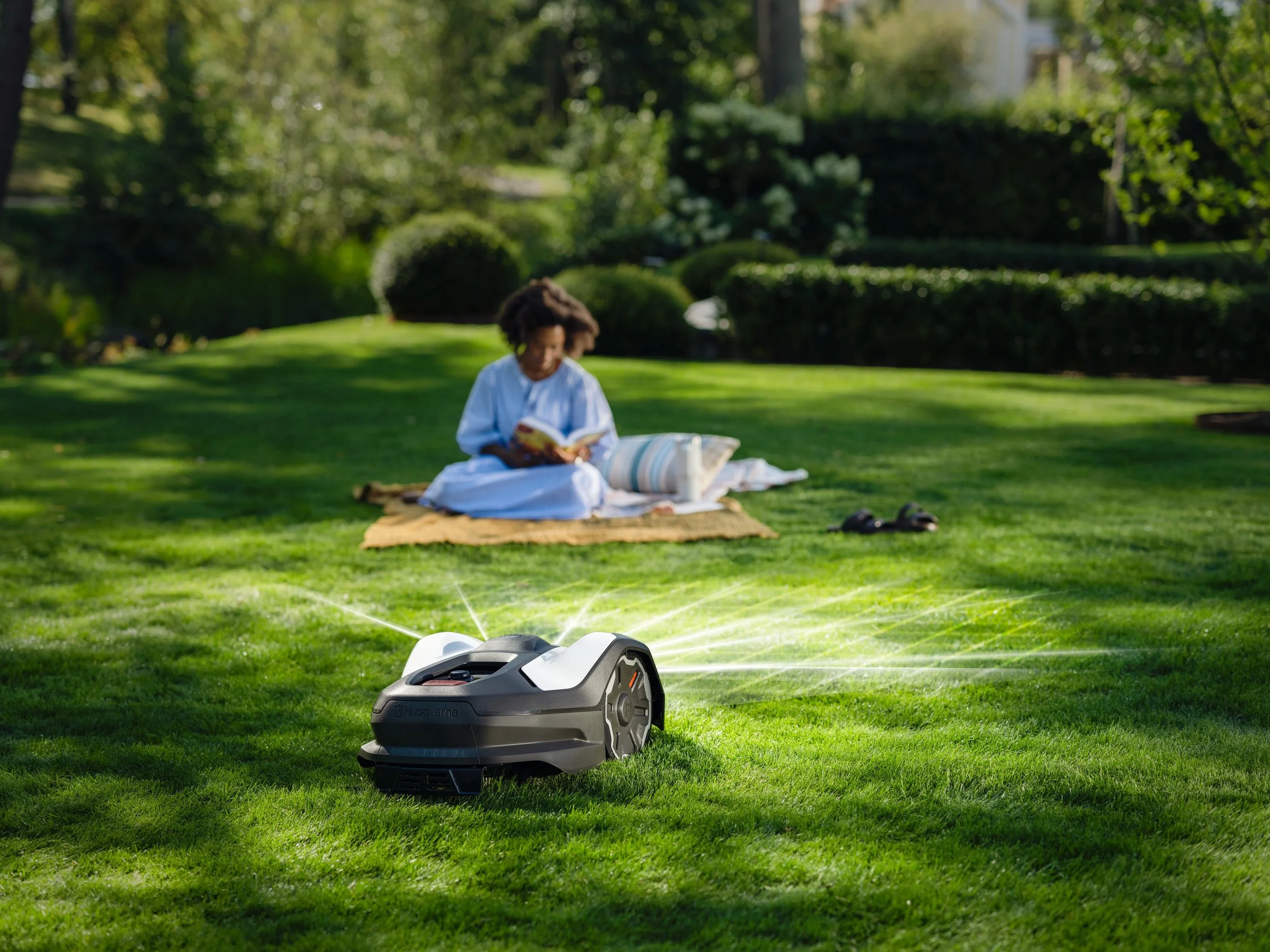 A person sitting on a blanket in a park reading a book, with a robotic lawn mower nearby spraying water on the grass, surrounded by trees and bushes.