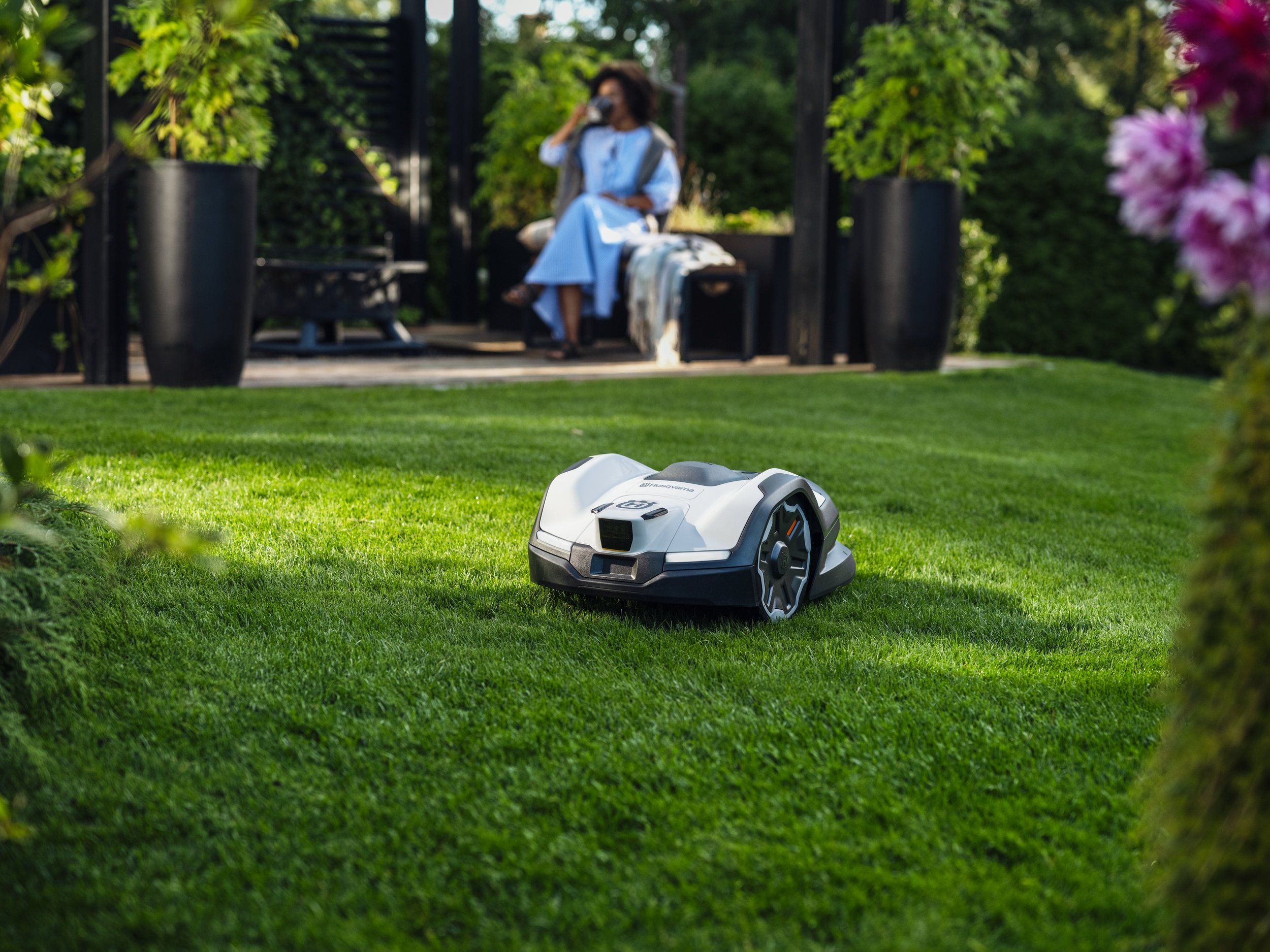 A robotic lawn mower on a well-maintained lawn with a woman sitting on a bench in the background, talking on a mobile phone in a garden setting.