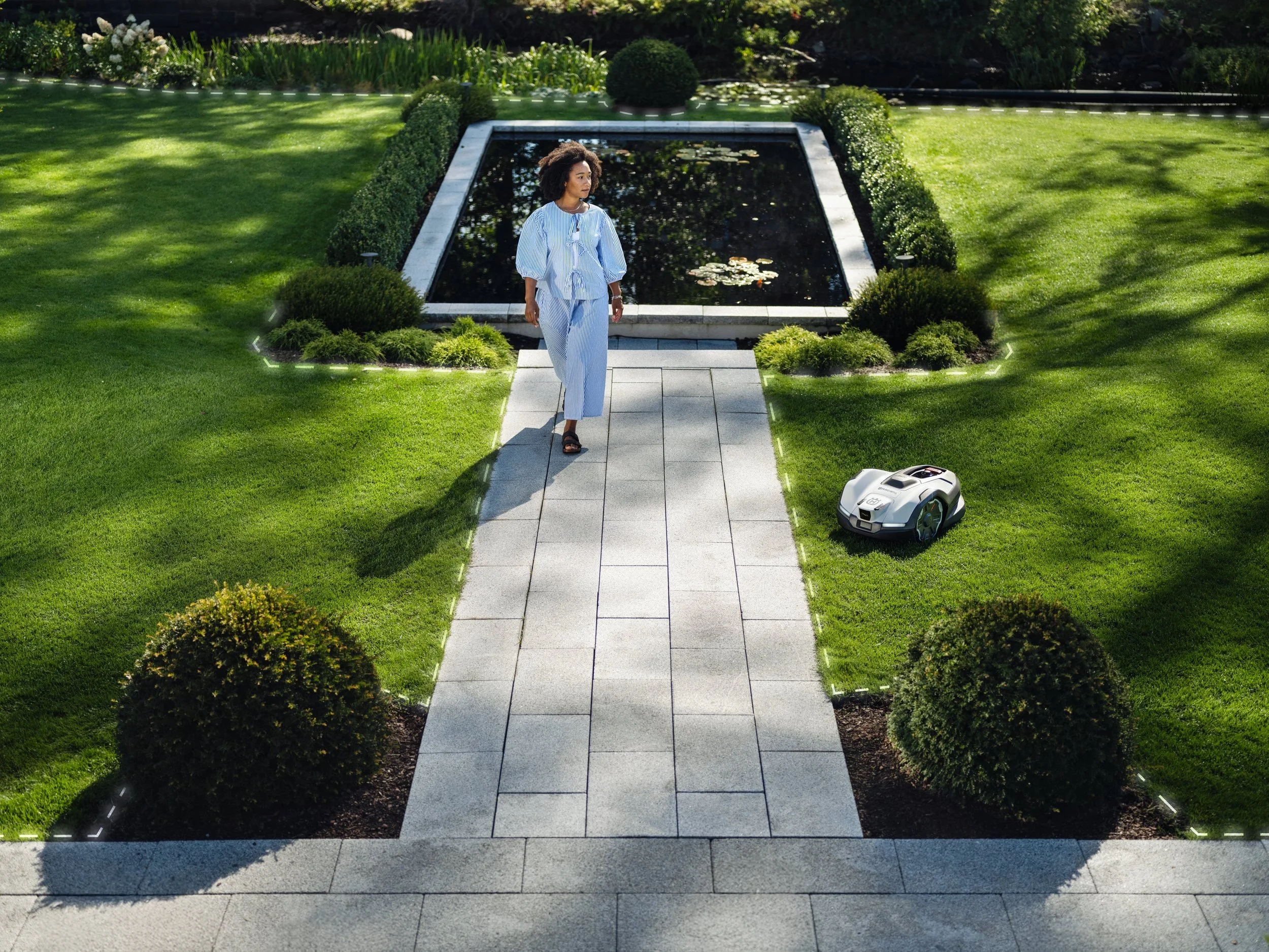 A woman in pajamas walking on a stone pathway in a well-maintained garden, with a robotic lawn mower nearby.
