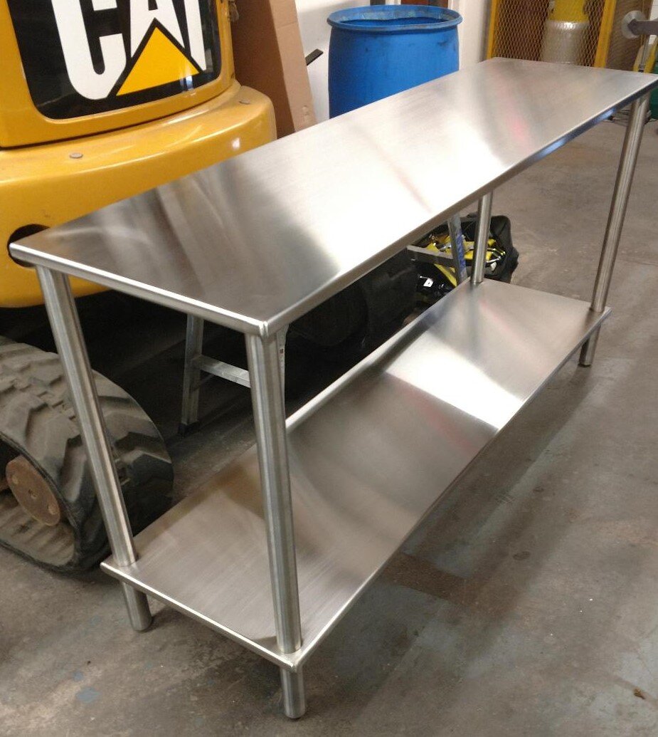 Stainless steel table with two shelves in a workshop setting next to a piece of industrial machinery.