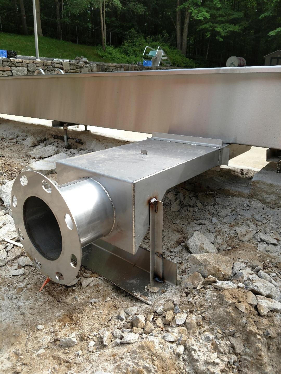 Metal exhaust vent installed on rocky ground outdoors near a stone wall.