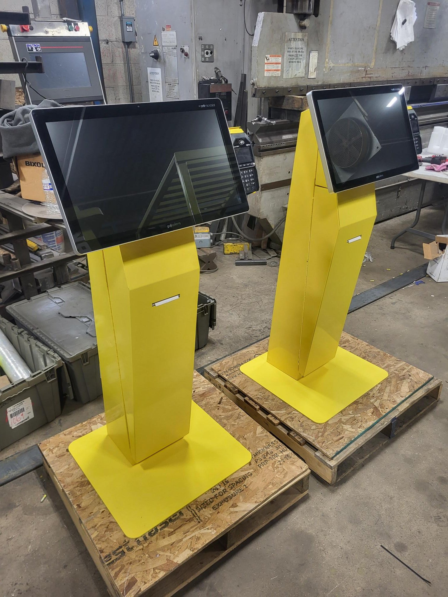 Two yellow interactive kiosks with touchscreens in a workshop setting, placed on wooden pallets.