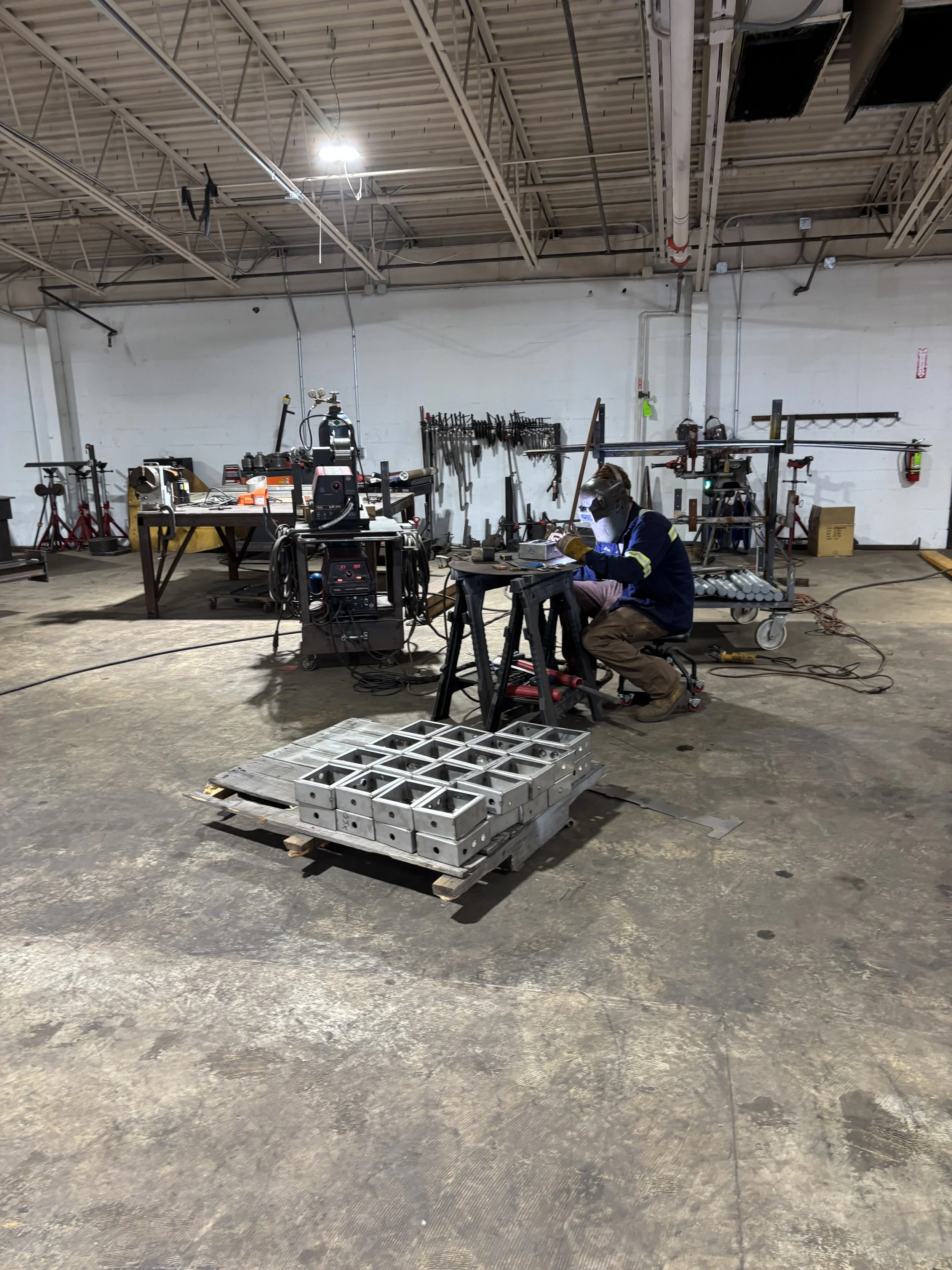 A worker welding or assembling metal parts at a workstation in a large industrial workshop.