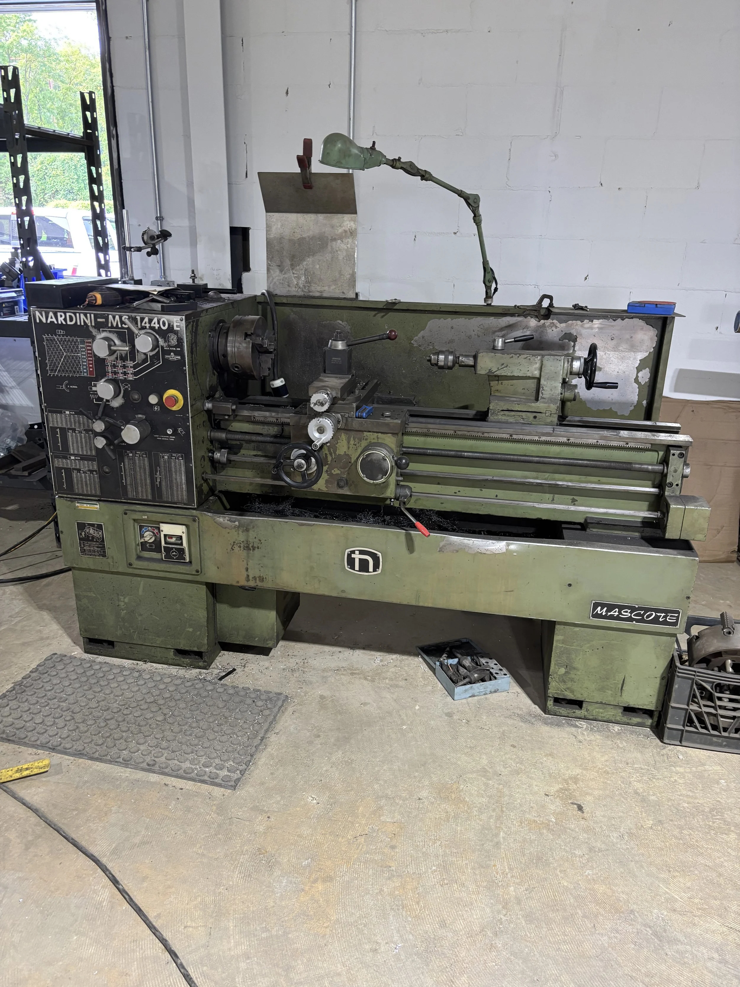 A vintage green metal lathe machine with controls and dials, situated in a workshop with concrete floor and white brick wall background.