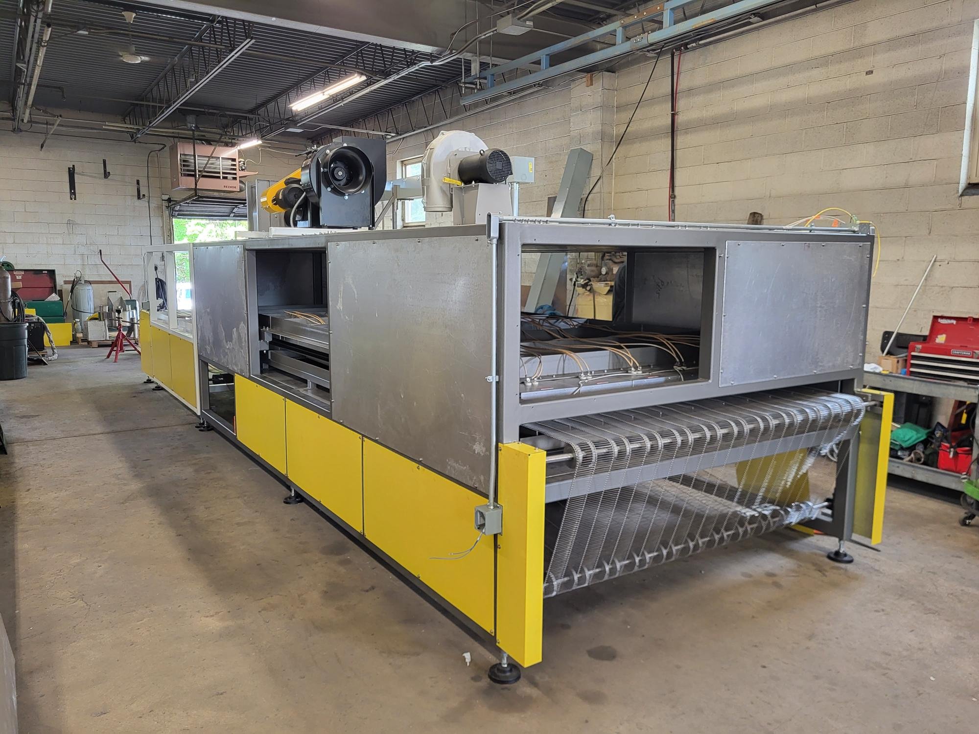 Industrial machinery in a workshop, featuring a large metal and yellow conveyor machine with rollers and fans, surrounded by tools and equipment.