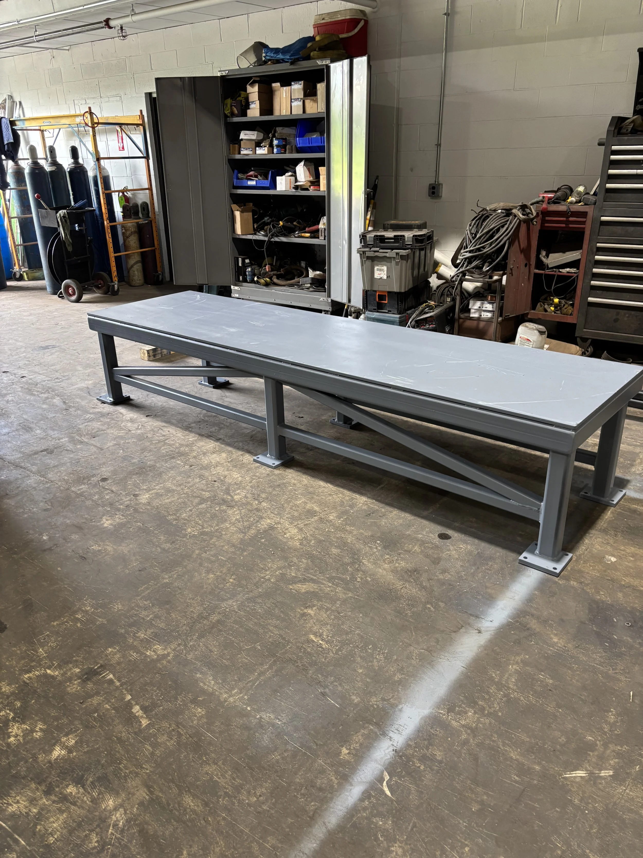 A metal workbench with a flat surface in a workshop or garage setting, with various tools, storage cabinets, and equipment in the background.