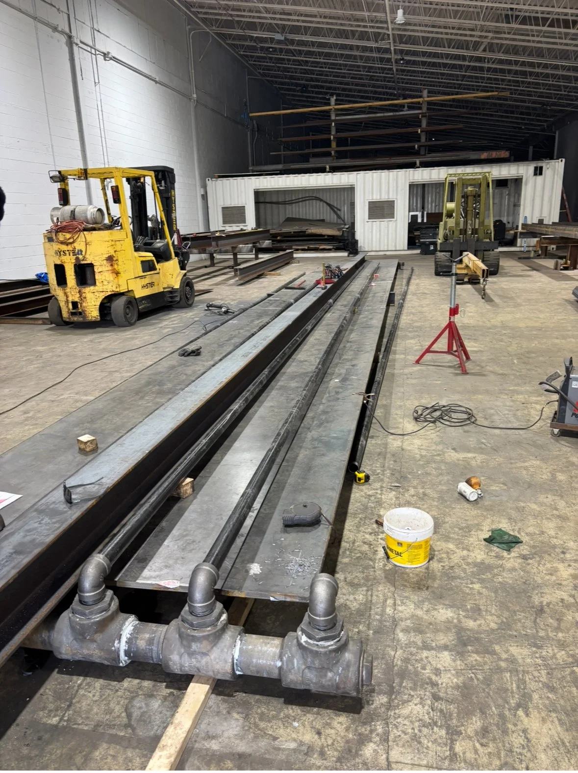 Interior view of a construction site showing steel beams, pipes, and construction equipment including a yellow forklift and a small yellow scissor lift in a large industrial space.