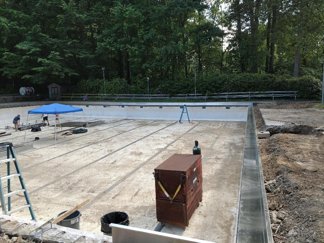 Empty swimming pool under construction with a blue canopy and ladders, surrounded by trees and seating.