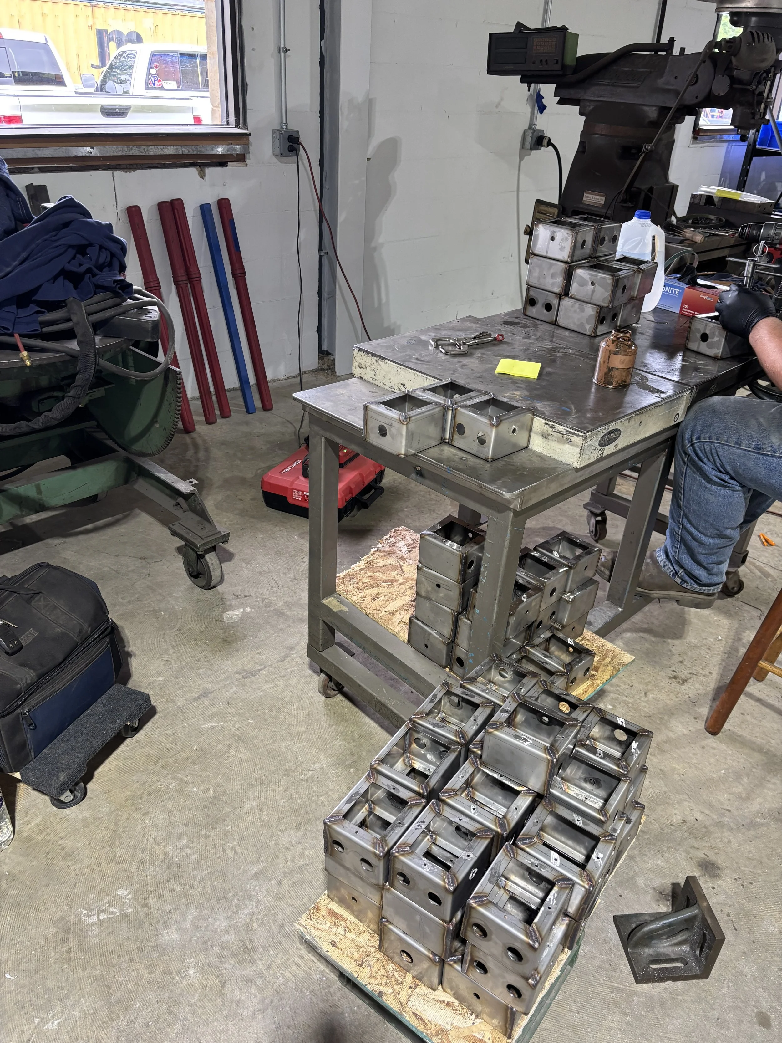 A workshop with a metal worktable holding several metal brackets and blocks, with more brackets stacked underneath, and a person working on the right side. There is a window with outside view, a cart with tools and a bag on the left, and various work
